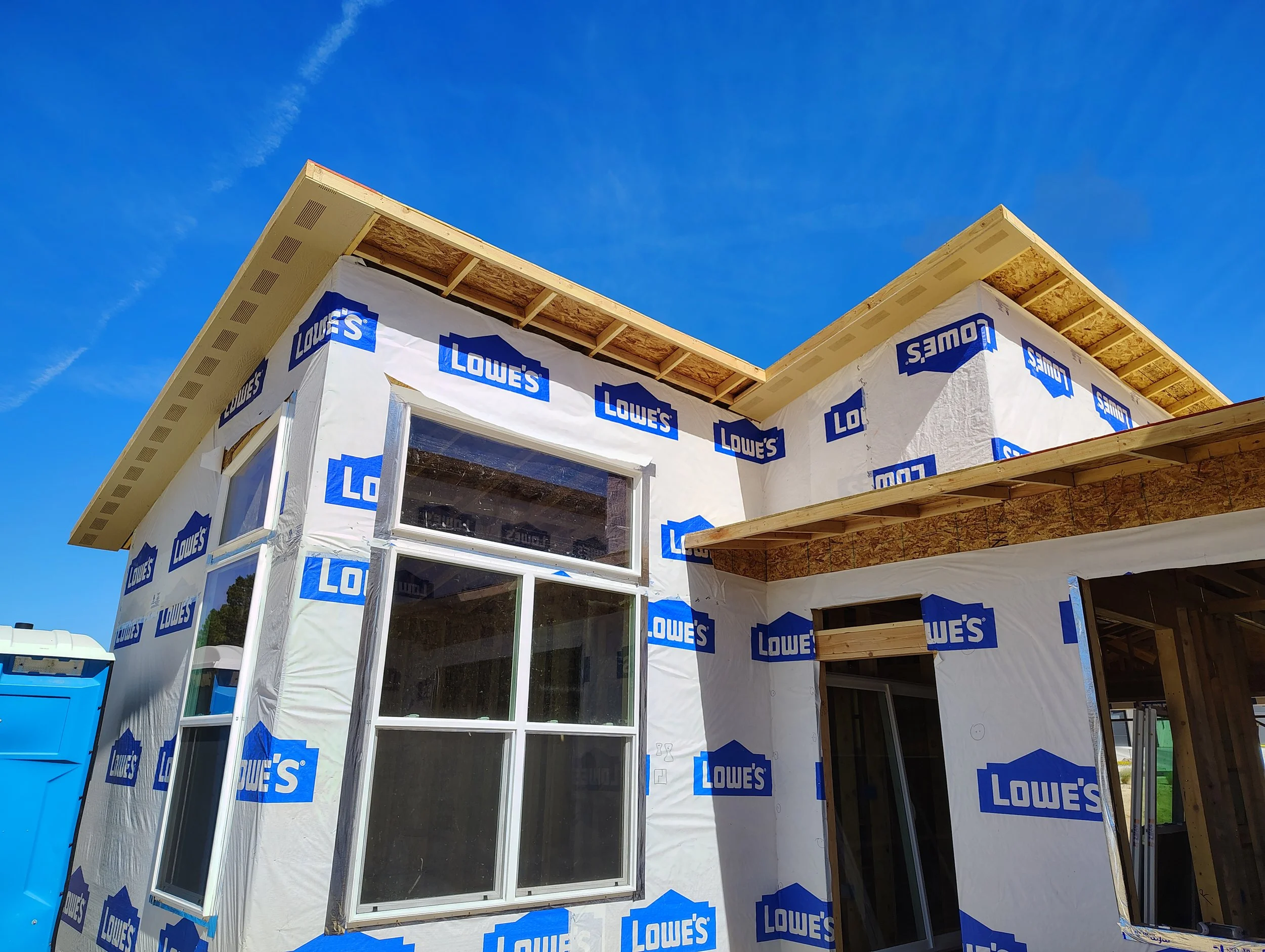 A house under construction with new framing, wrapped in white Lowe's building wrap, and a clear blue sky above.