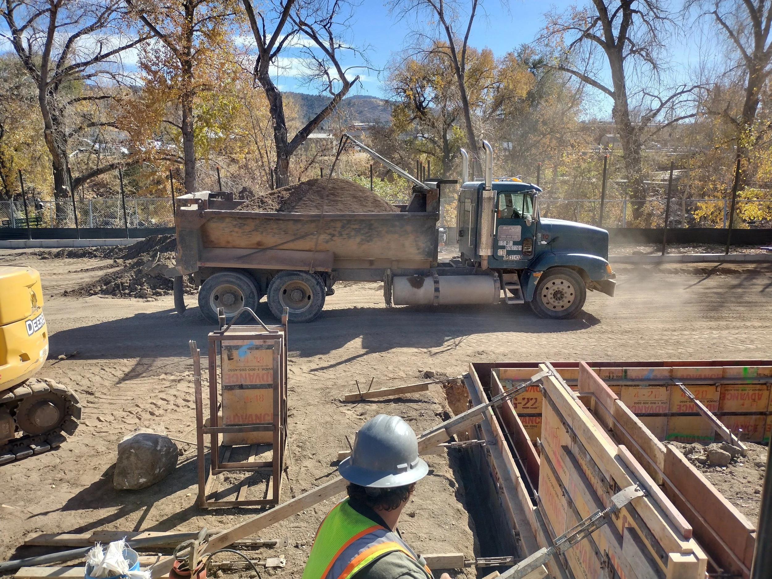 Construction site with a dump truck carrying dirt, construction worker wearing a white helmet and safety vest, trees with fall foliage in the background.