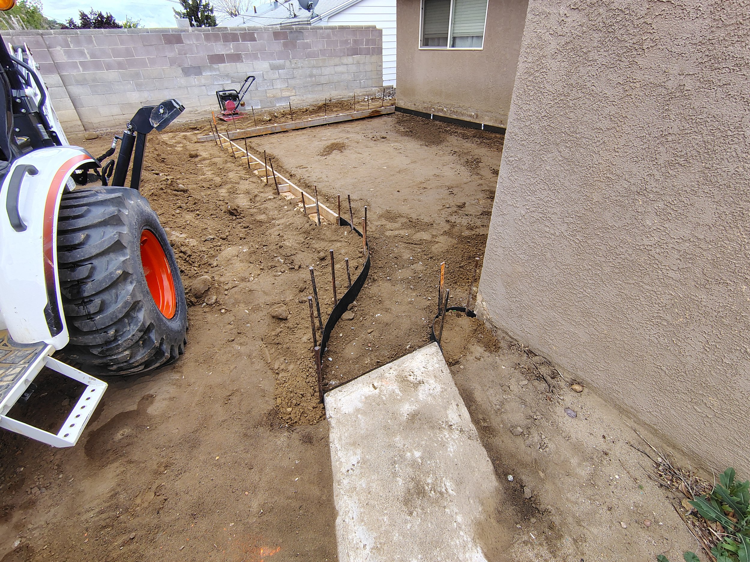 A construction site with exposed soil, wooden and metal framing, and a compacting machine, next to a house with a stucco exterior wall and a window, and a concrete block wall in the background.