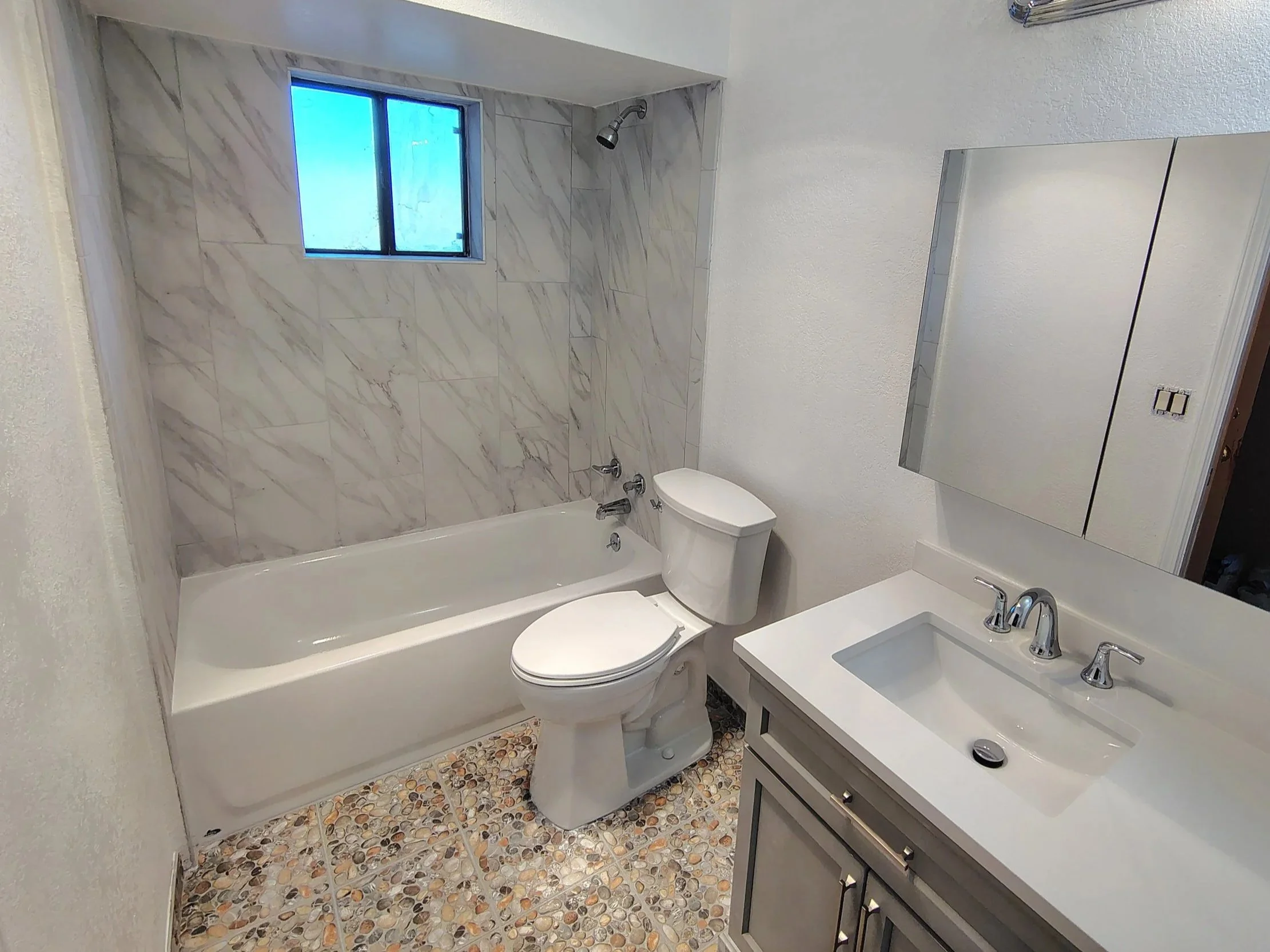 A small bathroom with a bathtub, toilet, and sink with a mirror. The wall above the bathtub is tiled with marble tiles. There is a small window above the bathtub, and a mirror on the wall above the sink. The floor has colorful pebble-like tiles.