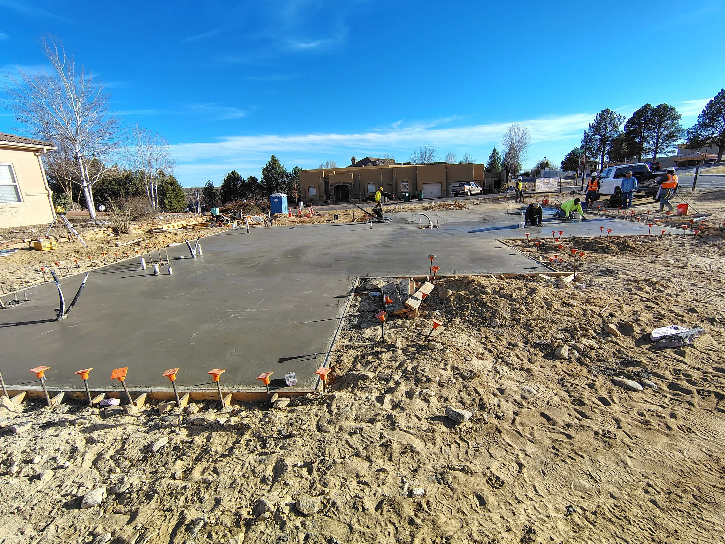 Construction workers are pouring and finishing concrete on a building site, with some workers in high-visibility vests and hard hats, and construction equipment and vehicles in the background under a clear blue sky.