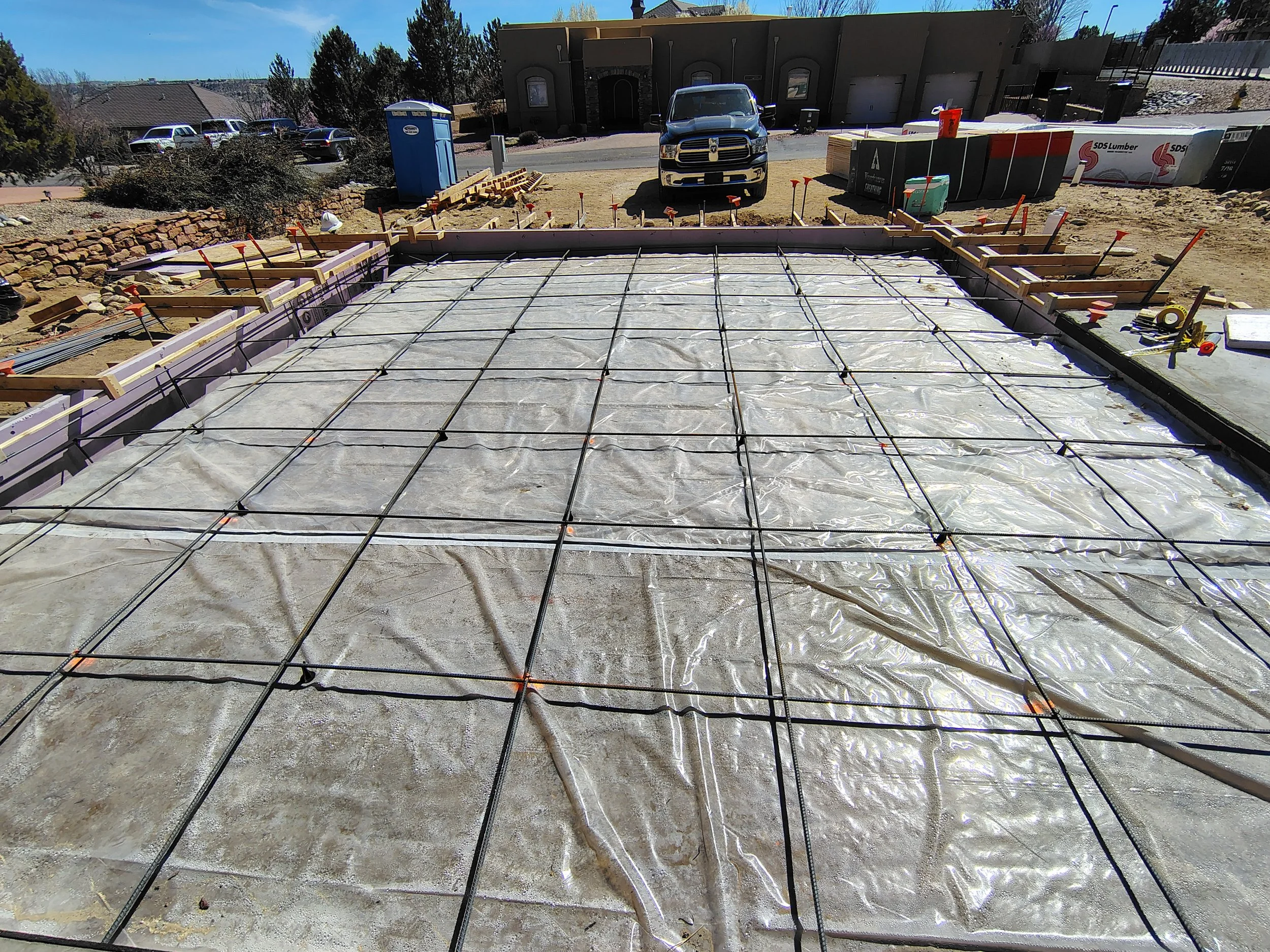 Construction site with a poured concrete slab reinforced with rebar, surrounded by wooden forms, in a residential area with parked cars, trees, a portable toilet, and construction materials in the background.