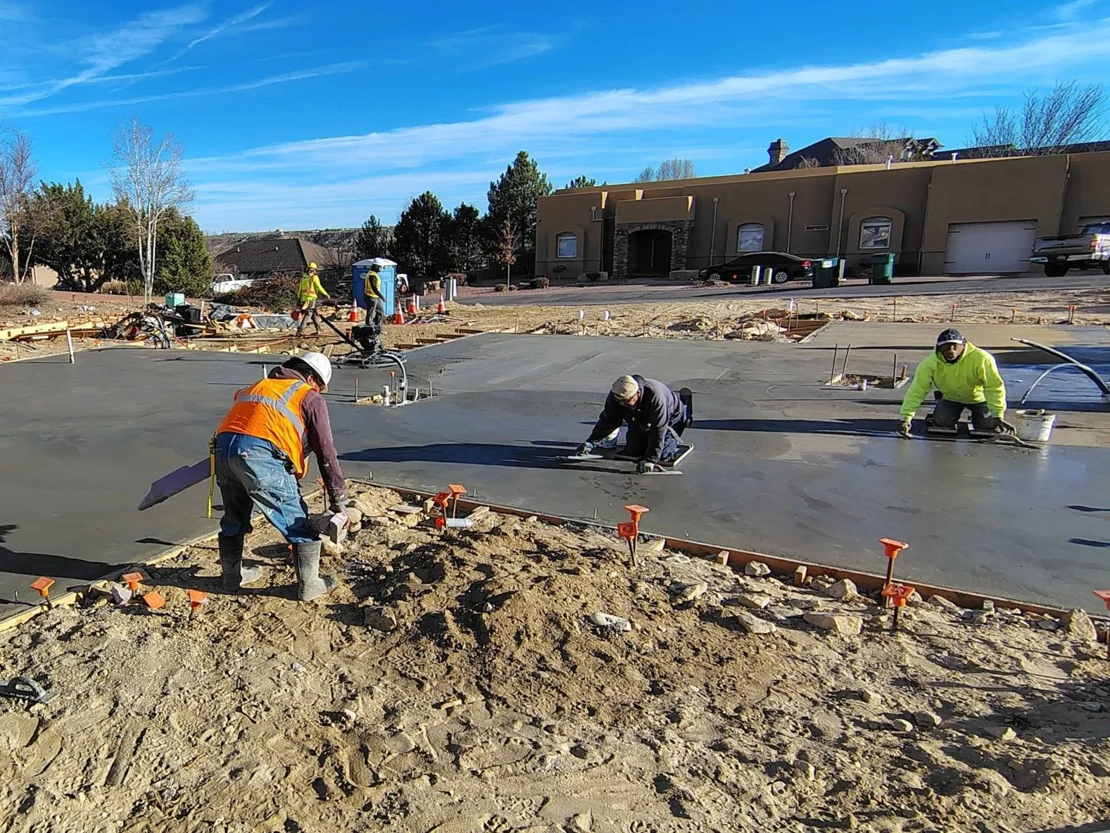 Construction workers pouring and smoothing concrete on a building site with an apartment building in the background and clear blue sky.