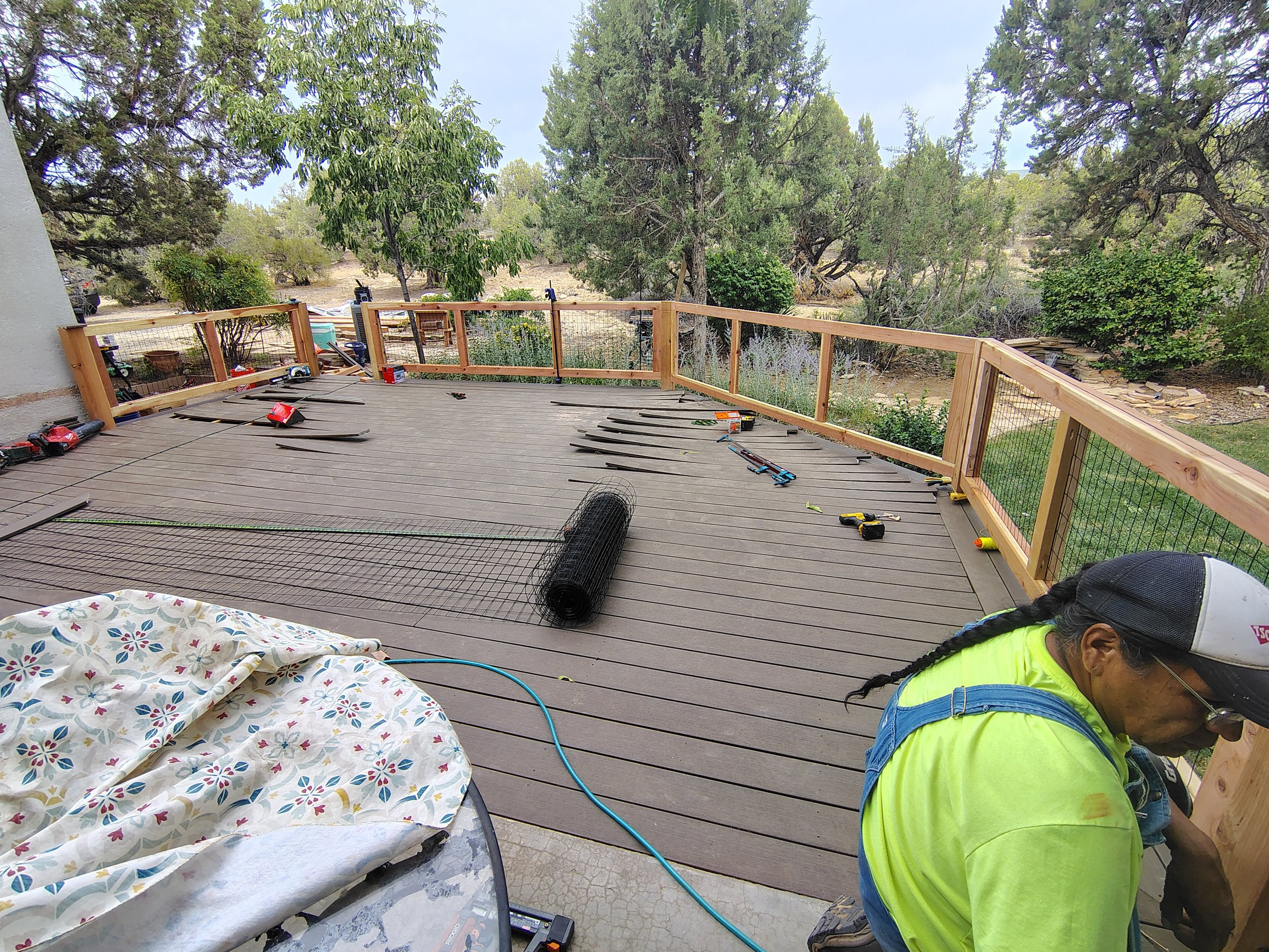 Workers are installing a wooden deck, with tools and materials scattered around, in a backyard with trees and bushes.