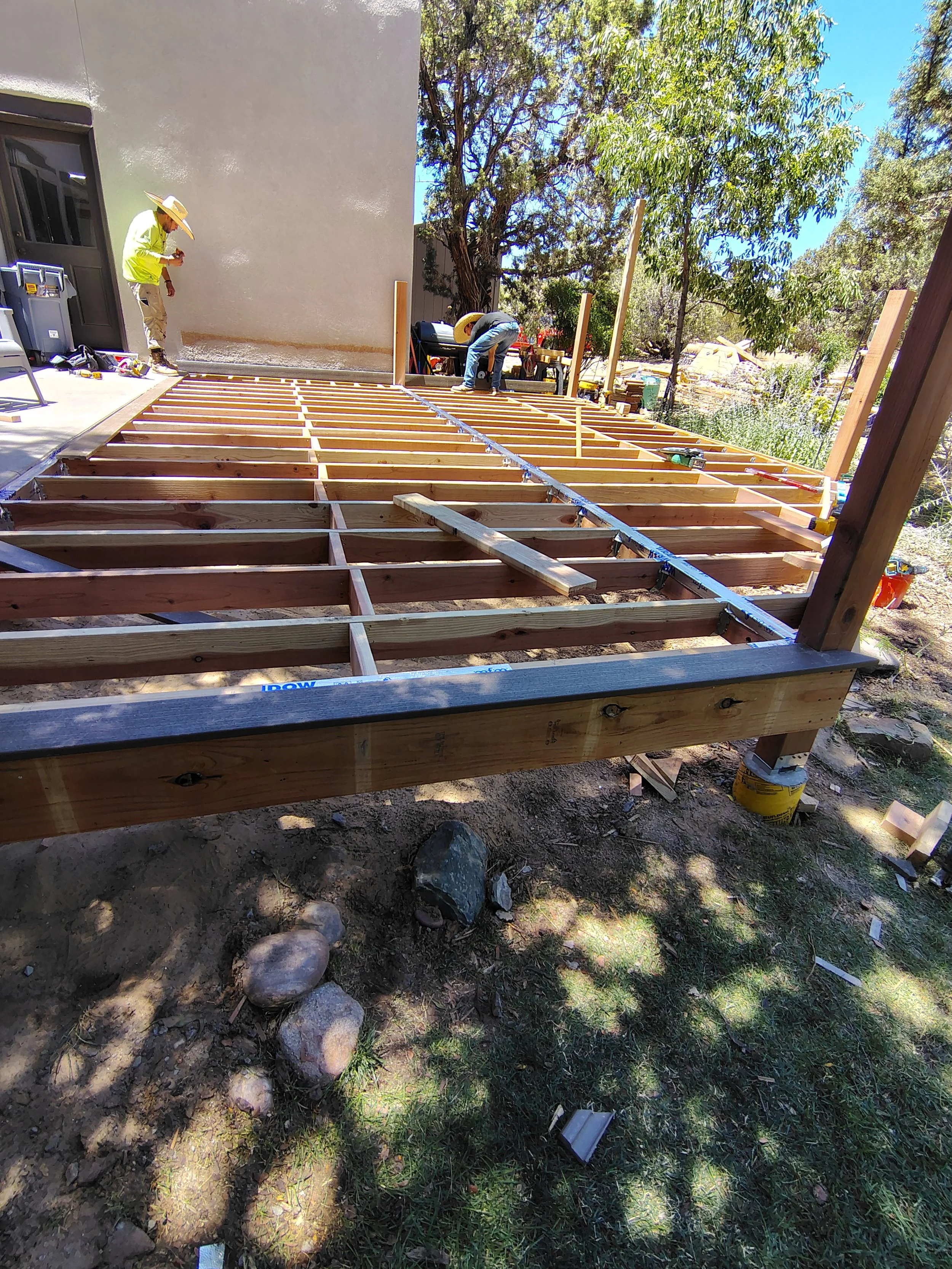 Two workers are constructing a wooden deck outside a house, with one worker cutting wood and the other also working on the deck, surrounded by tools and materials, with trees in the background.