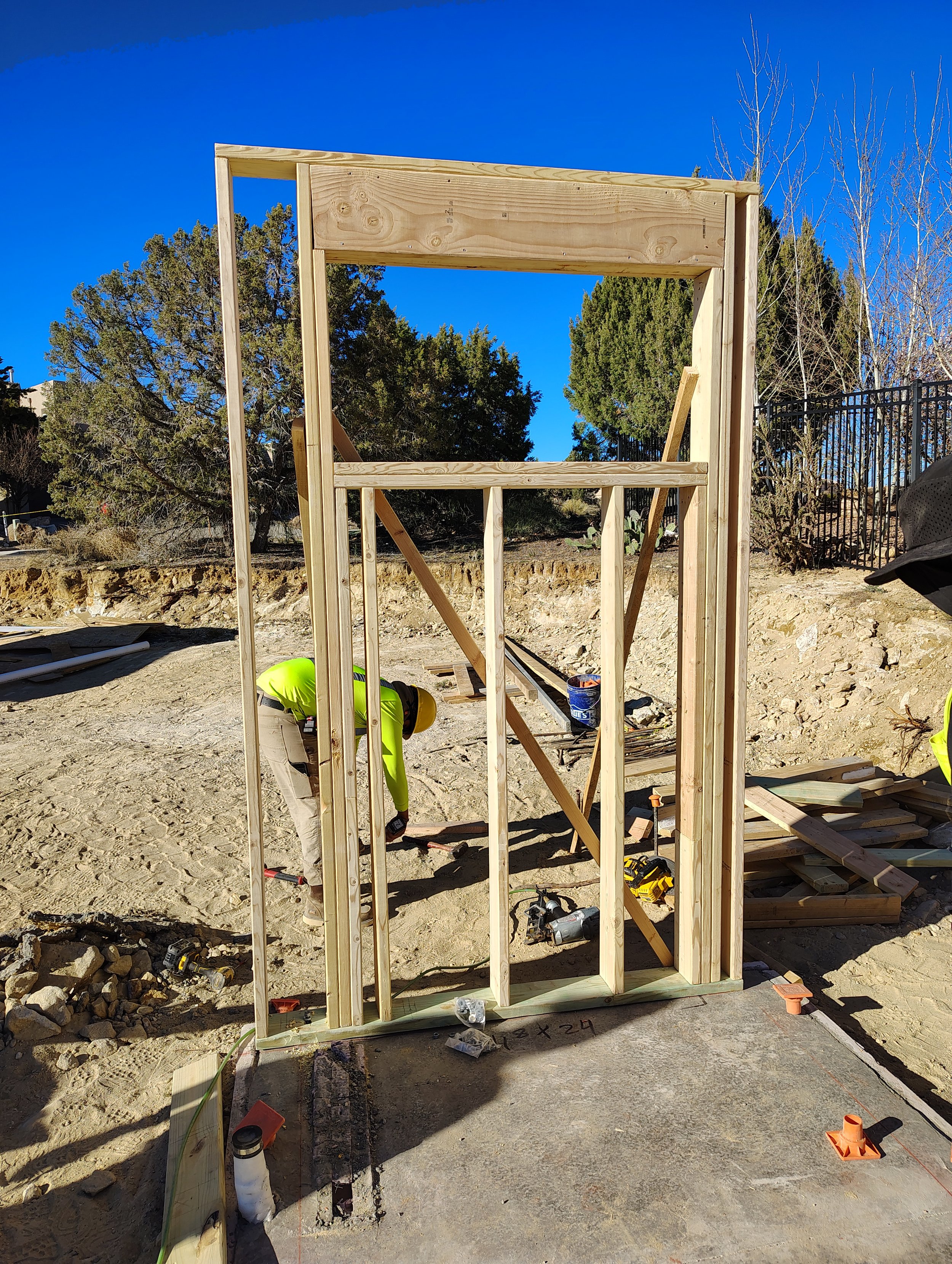Construction site with wooden framing for a small structure, worker in yellow safety vest and helmet below, tools and construction materials scattered around.