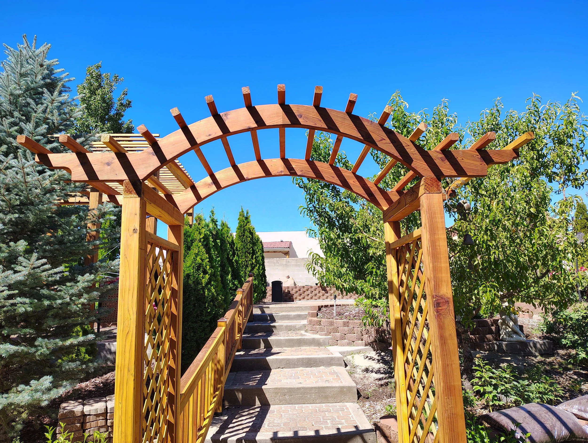Wooden garden arched trellis pathway with brick steps and lush green trees and shrubs under a clear blue sky.