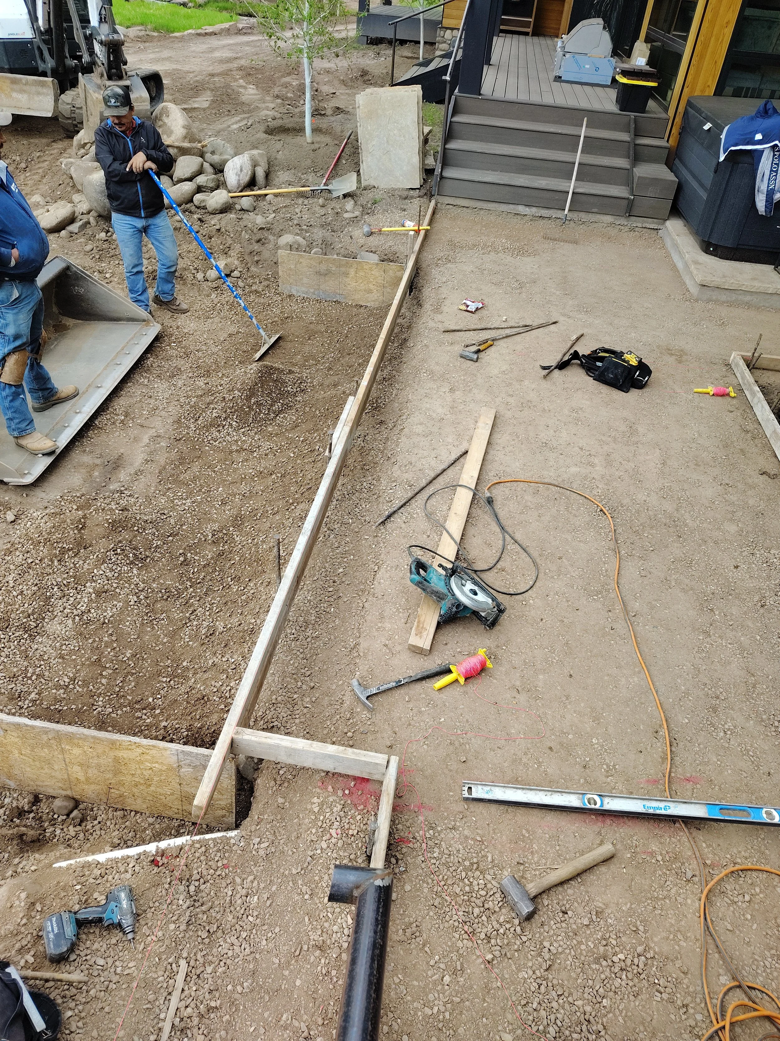 Construction site with workers and tools, preparing a foundation for a building or structure. Wooden framing, tools, and gravel visible.