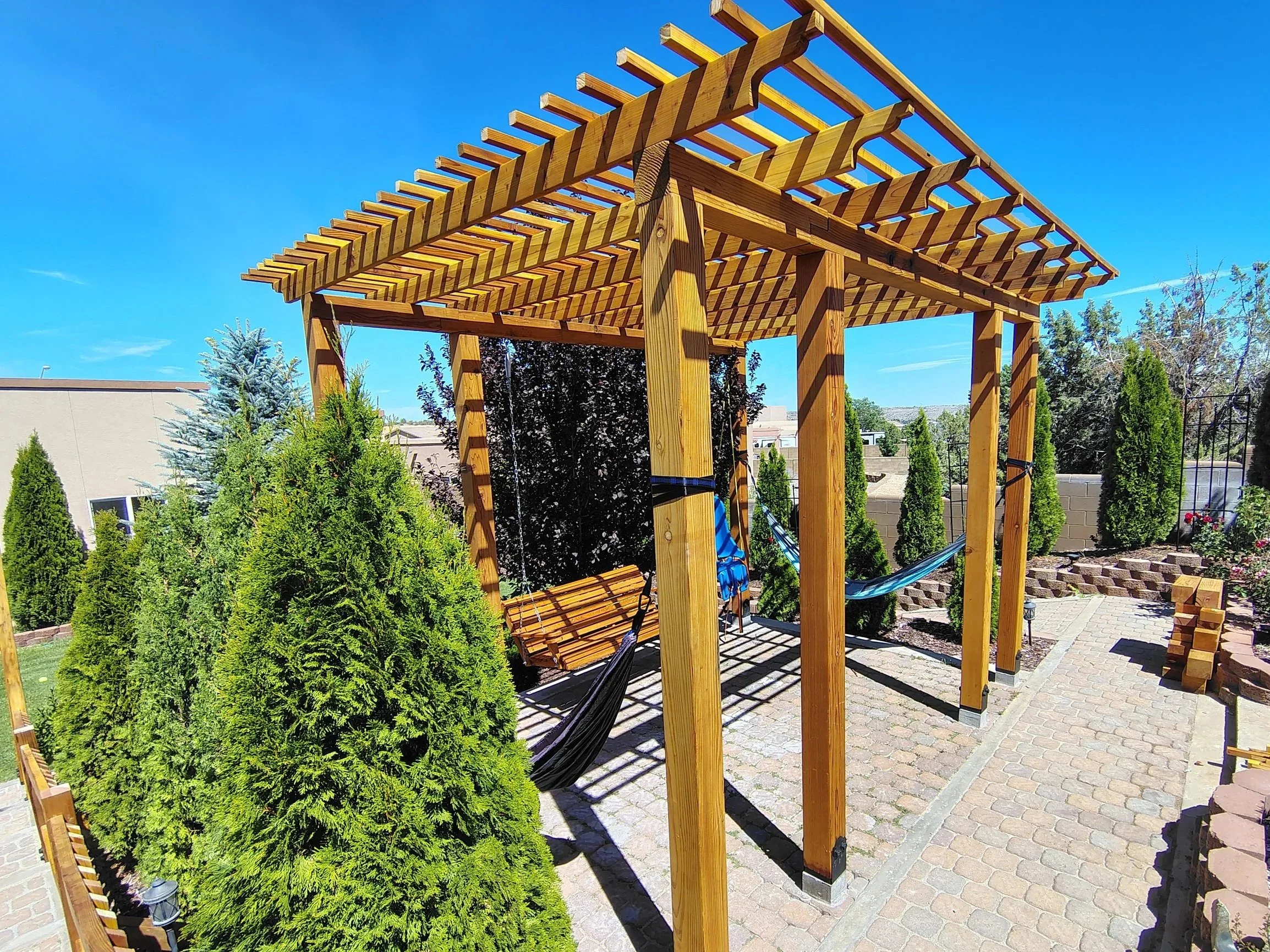 A partially constructed wooden pergola with a swing hanging underneath, surrounded by green bushes and trees in a backyard with paving stones.