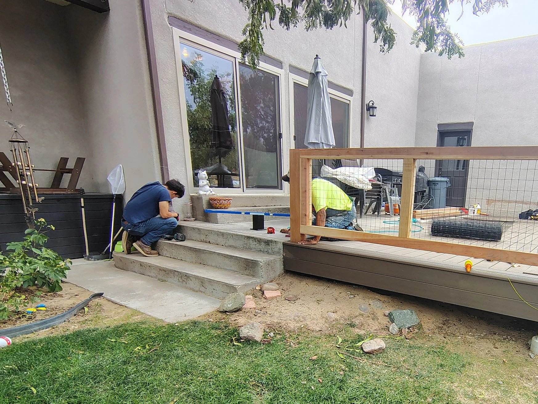 Two men are working on a home renovation project on the back porch of a house, with tools and construction materials scattered around. One man is crouched near the stairs checking his phone, while the other is kneeling on the deck, working with his hands. There are sliding glass doors, a fence, and outdoor furniture in the background.