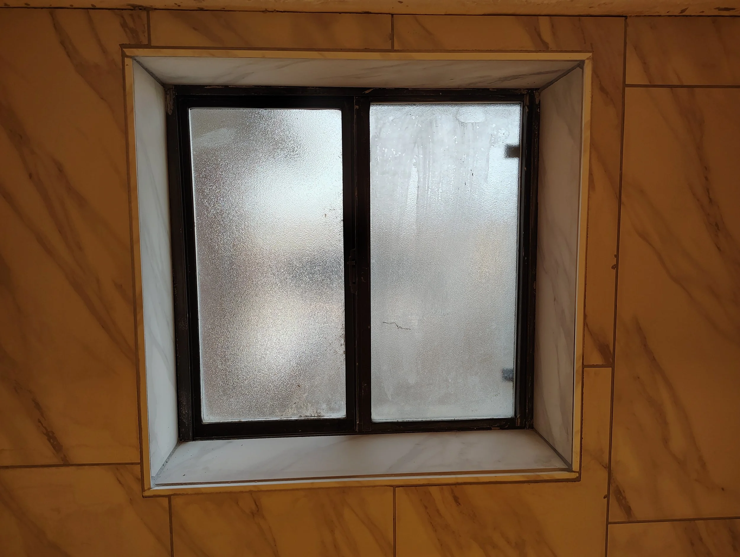 View of a small window with frosted glass, set in a wall with brown and beige tiles.