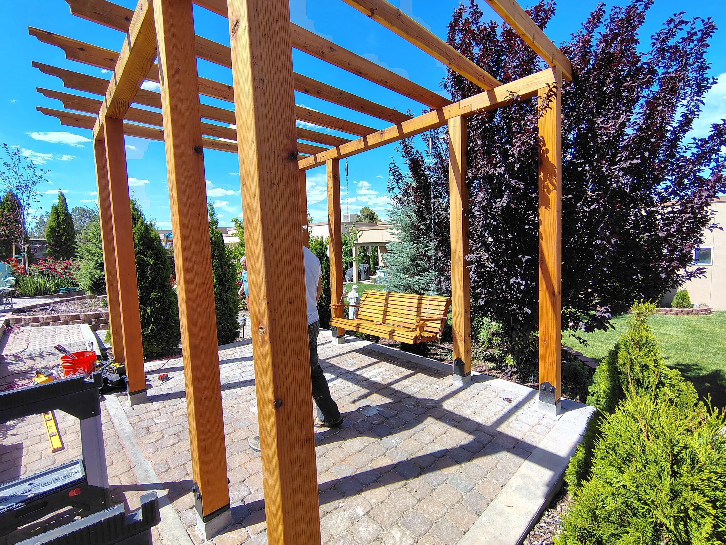 The image shows a wooden pergola under construction in a backyard garden with a bright blue sky and lush trees and plants surrounding the area.
