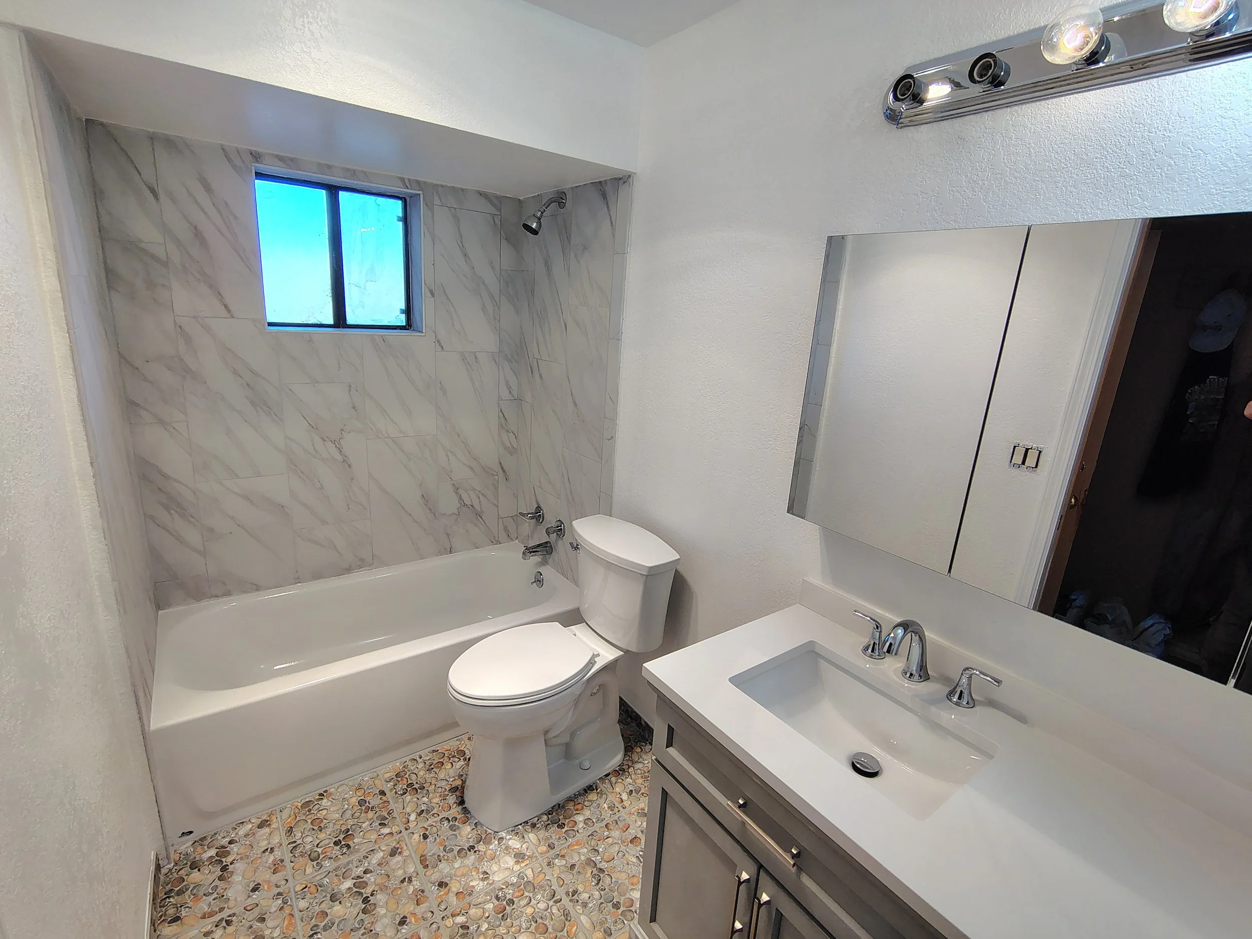 A small bathroom featuring a bathtub with a marble tiled wall and shower head, a toilet, a vanity with a sink, a mirror, and a window with a blue sky visible outside.