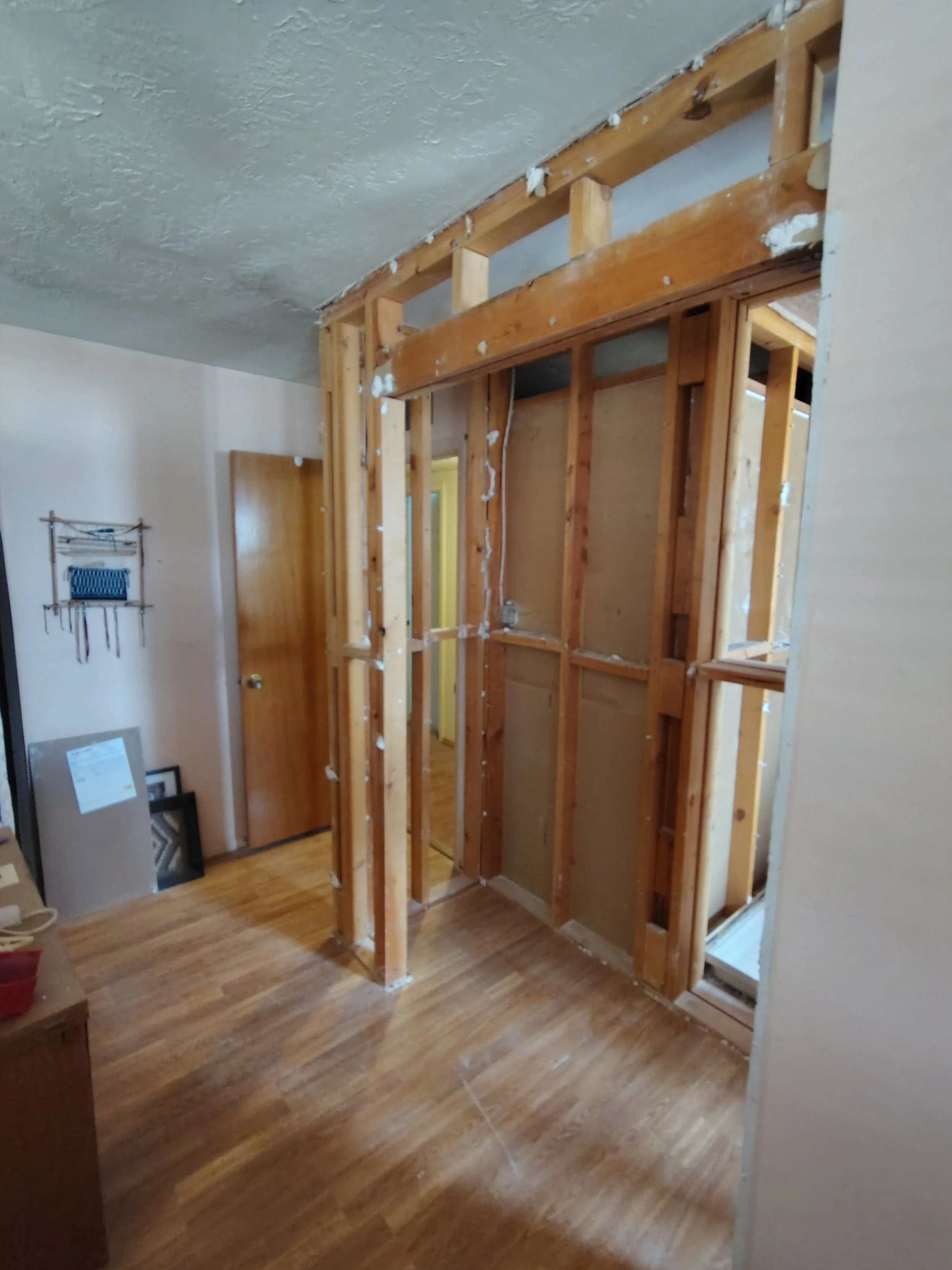 Unfinished interior room showing wooden framing for a wall or doorway under construction, with some tools and picture frames on the floor and a wall with a small decorative hanging.