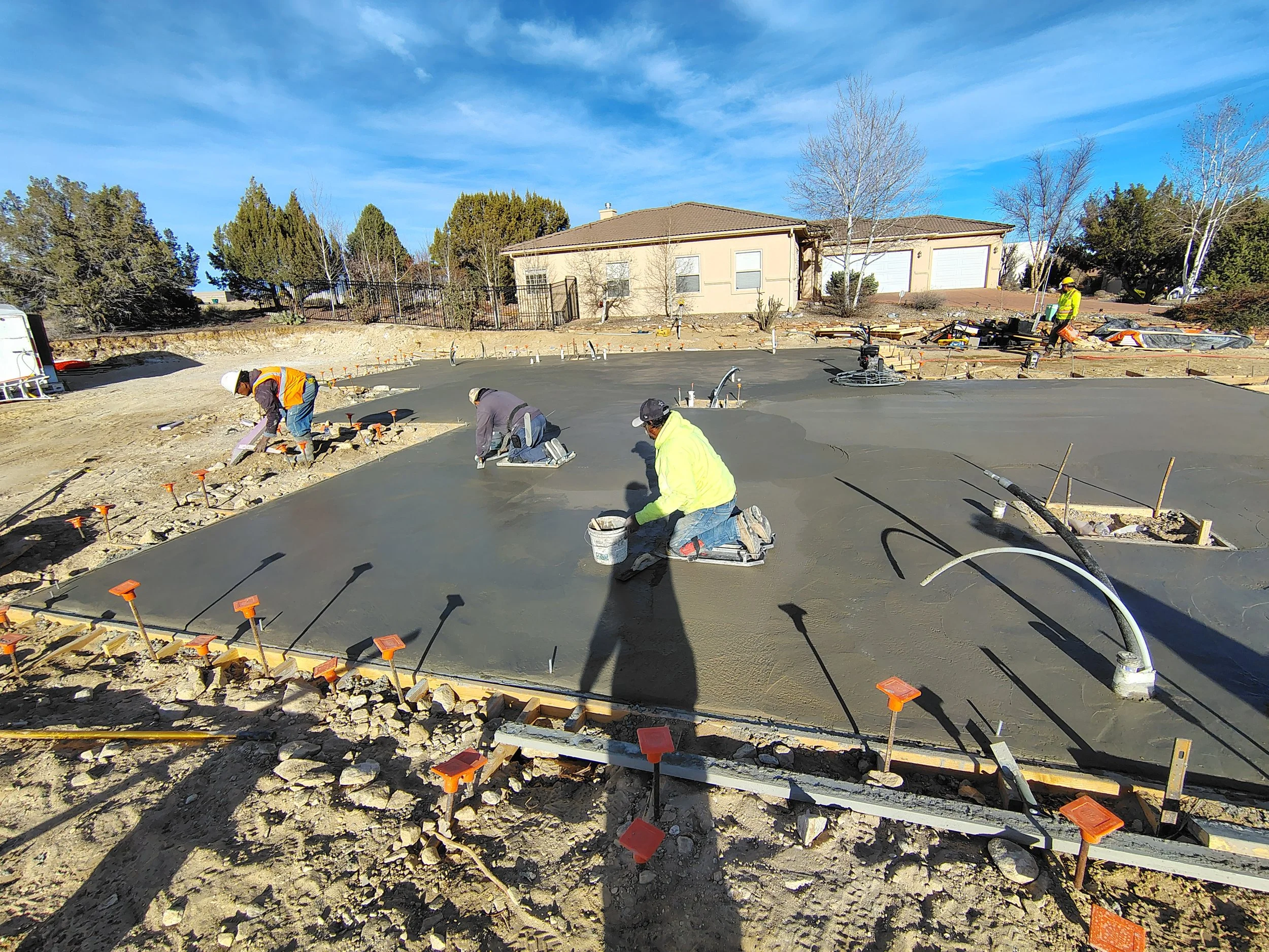 Construction workers pouring and smoothing concrete on a residential driveway in a suburban neighborhood with trees and a house in the background.