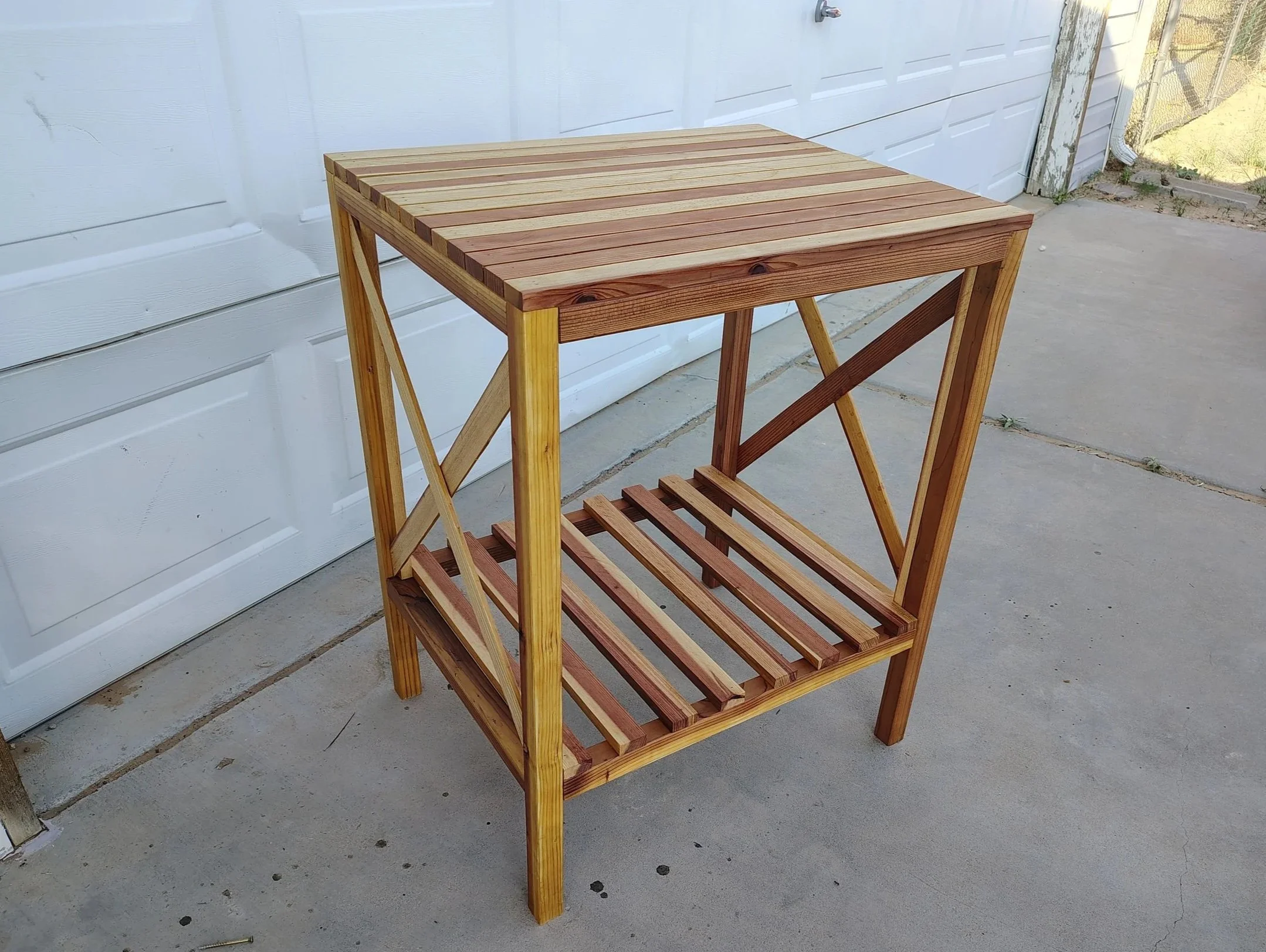 A wooden shelf with a slatted top and bottom shelf, placed on a concrete patio next to a white garage door.