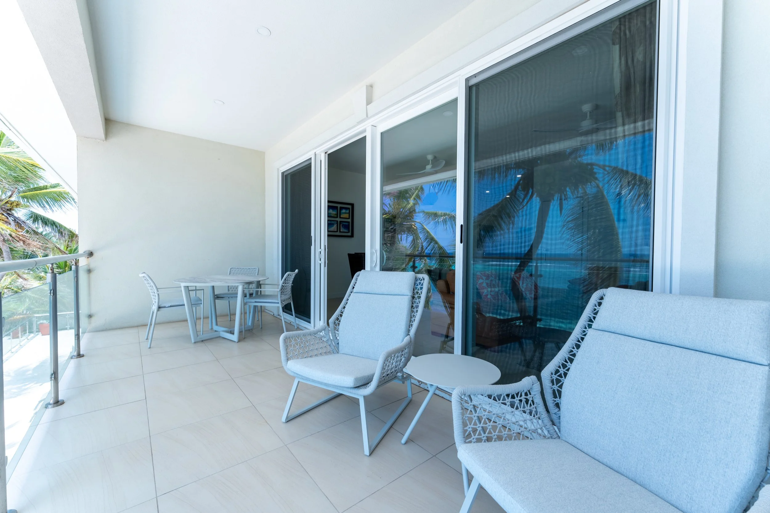 Balcony with white outdoor chairs, a small round table, and glass sliding doors reflecting palm trees and a blue sky.