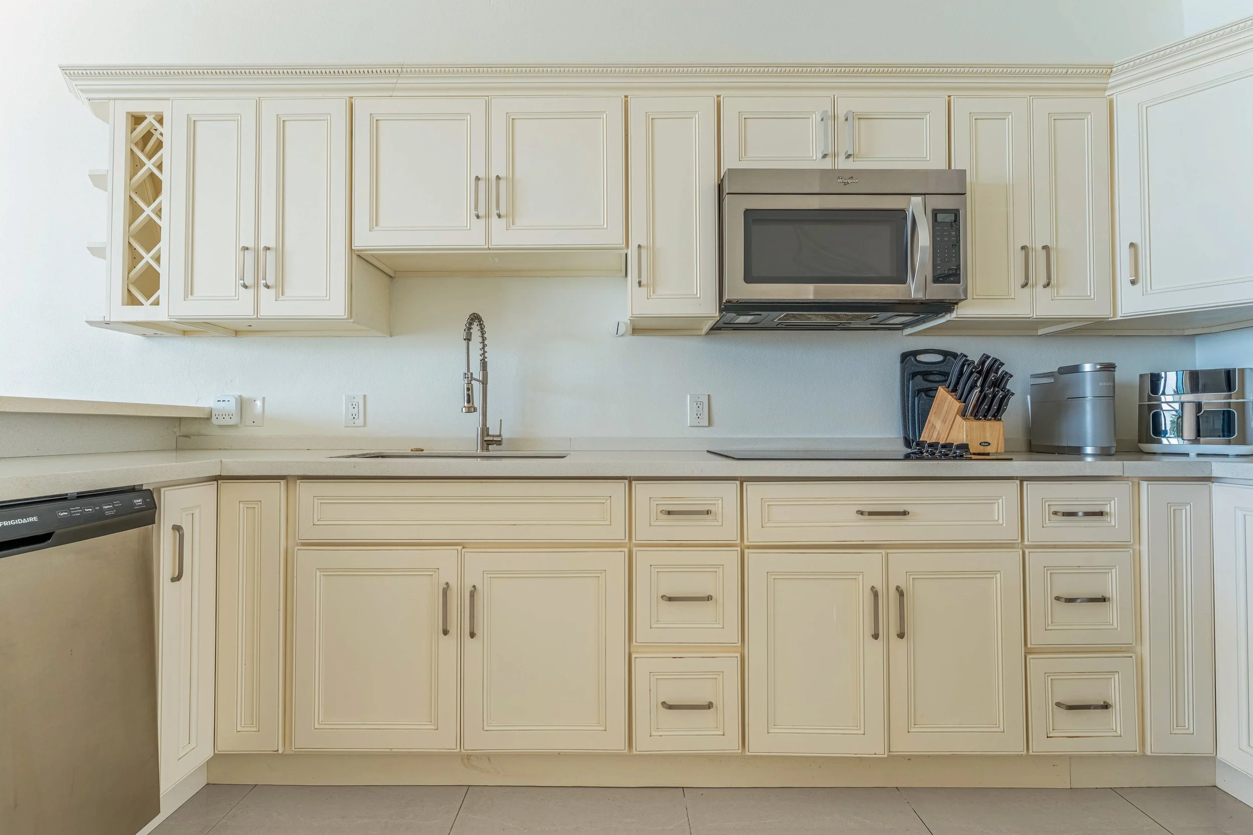 Kitchen with cream-colored cabinets, a stainless steel microwave above a stove, a sink with a spring coil faucet, a cutting board with knives, and appliances including a toaster and a trash can on the counter.