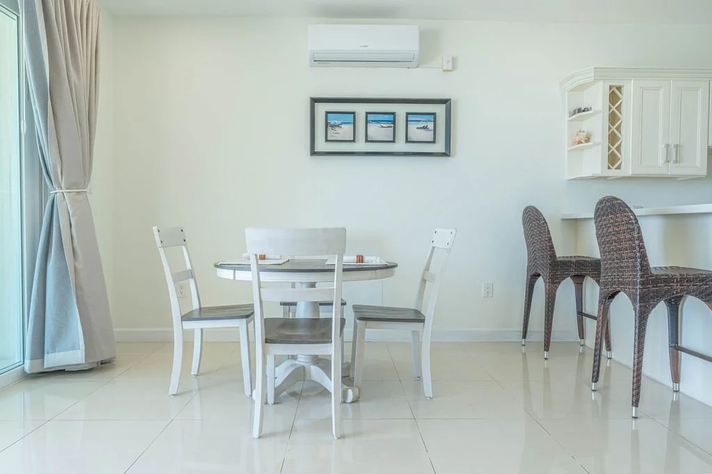 Dining area with white round table and four matching chairs, bar seating with two woven chairs, framed beach photos on the wall, air conditioning unit above the photos, and a door with curtains to the left.