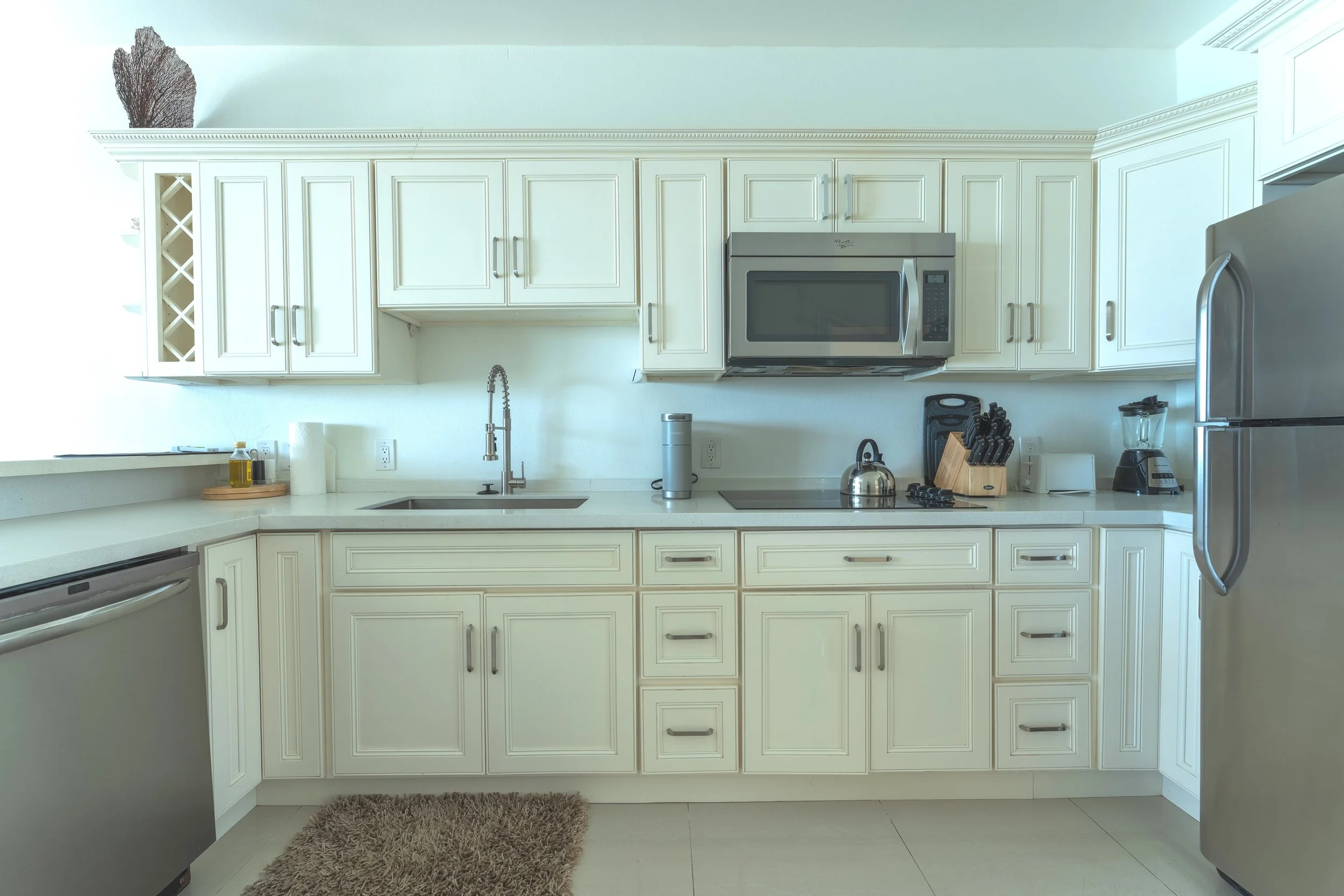 Kitchen with white cabinets, stainless steel microwave, and refrigerator, countertop with appliances including a blender and knife block, sink with a faucet, and a brown rug on the floor.