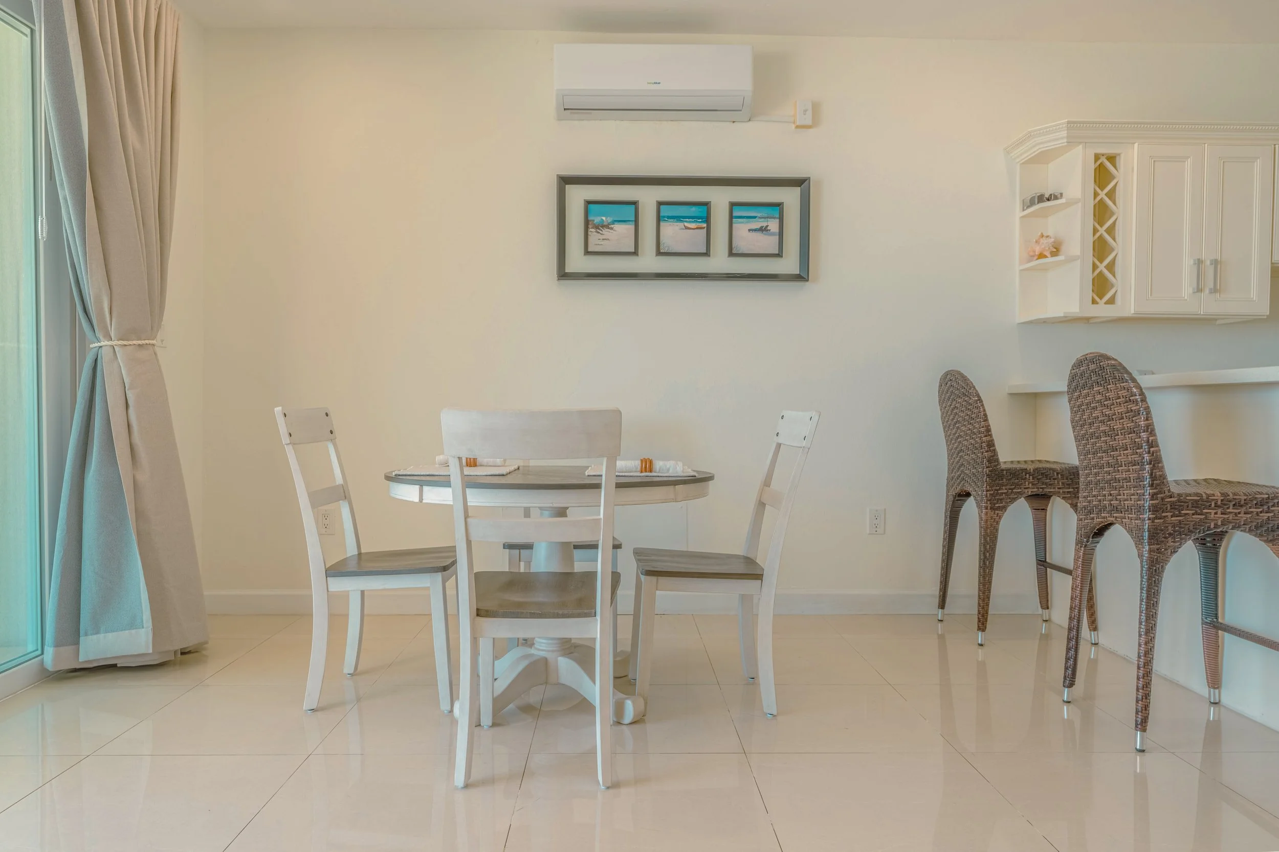A dining area with a round white table set with place mats, four white chairs, and two wicker bar stools. There is a sliding glass door with beige curtains on the left, a framed set of three beach-themed pictures on the wall, and a white cabinet with