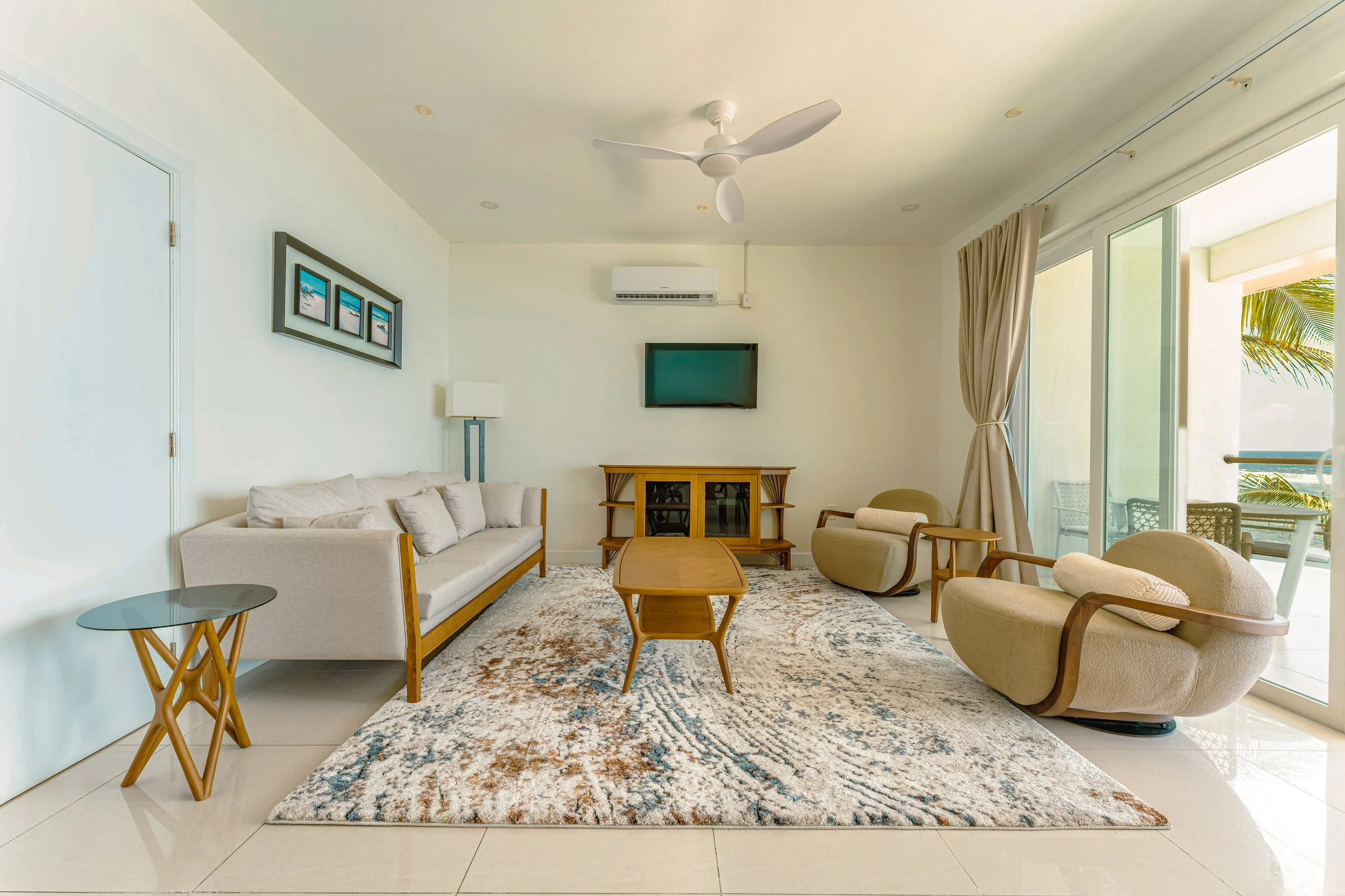 Living room with beige sofa, matching armchairs, wooden coffee table, area rug, wall-mounted TV, air conditioning unit, framed artwork, floor lamp, sliding glass door with curtains, and outdoor balcony with palm tree