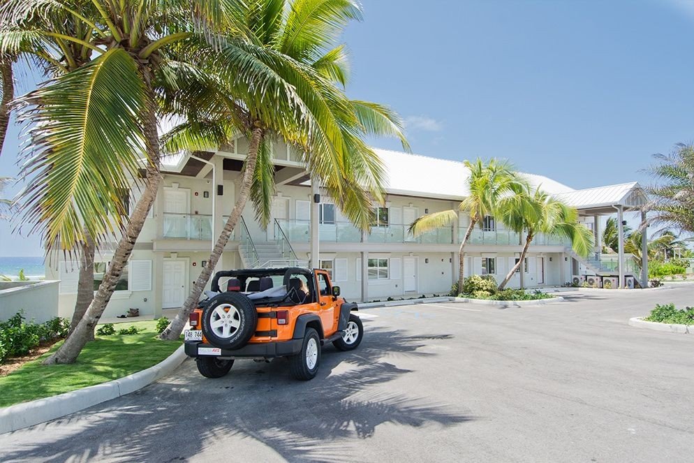 Beachside apartment building with palm trees, a bright orange Jeep, and parking lot.