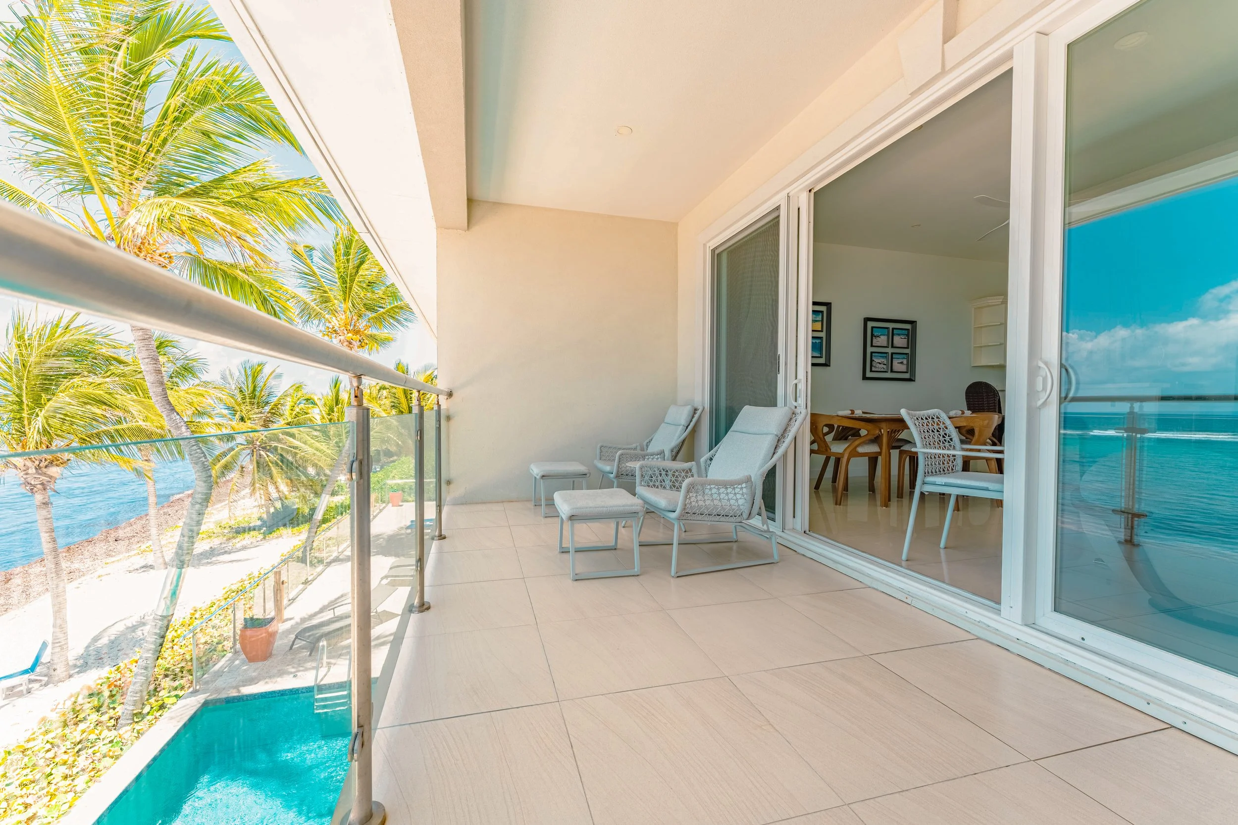 Balcony with white chairs overlooking palm trees, ocean view, and sliding glass door leading to interior dining area.
