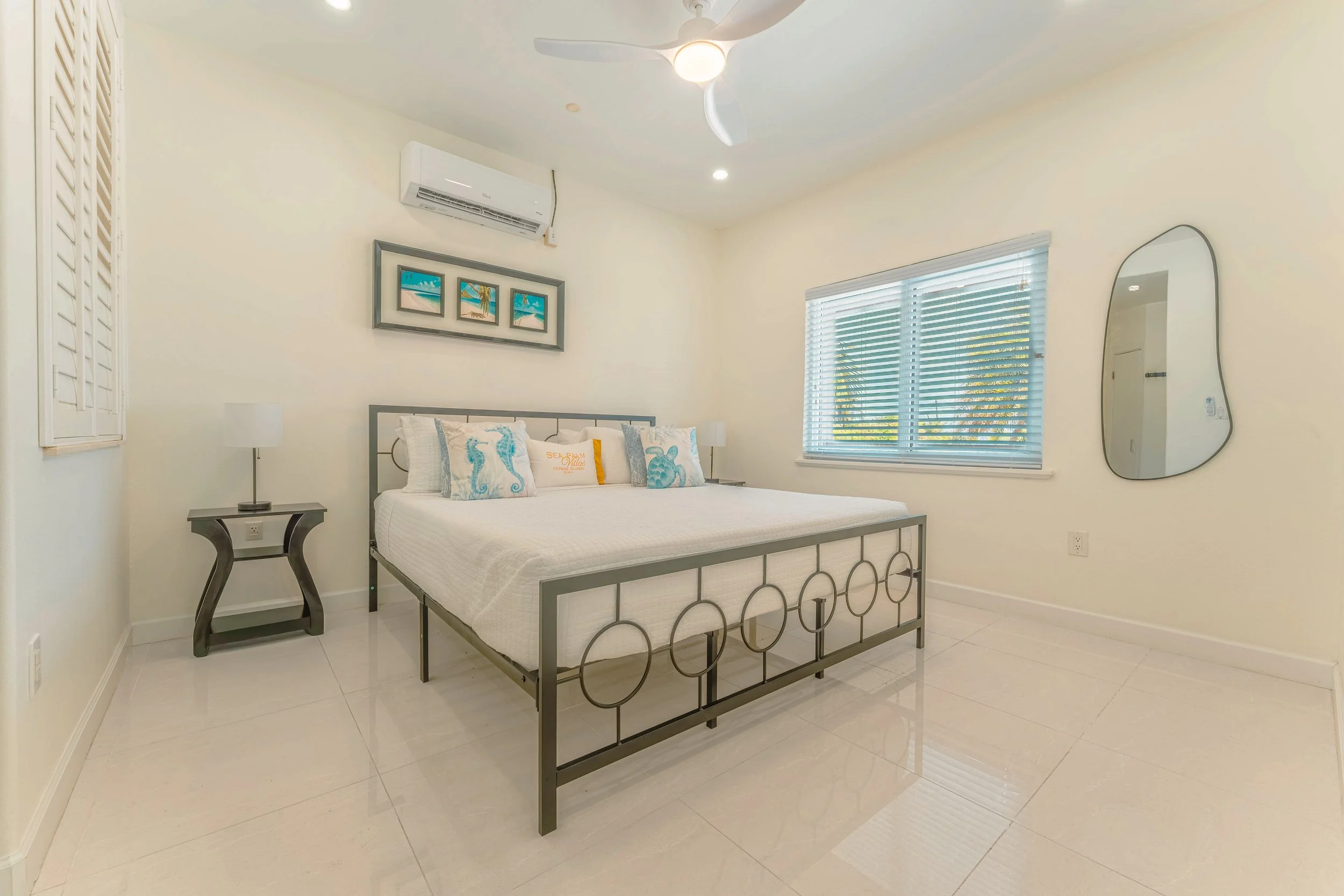 A bedroom with a white wall, large window with blinds, a bed with white bedding and three decorative pillows featuring sea horses, a modern bedside table with a white lamp, a split air conditioning unit above the bed, a minimalist mirror on the right