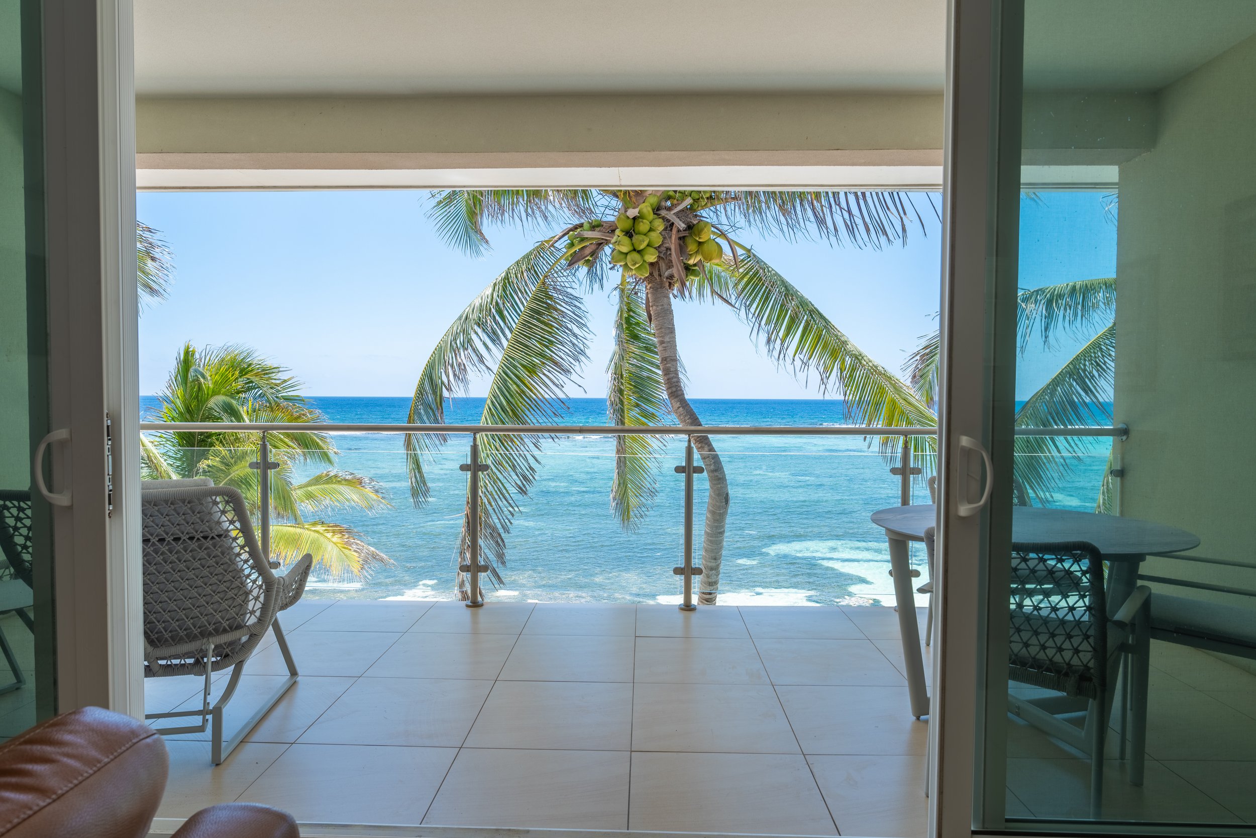 View from a balcony overlooking the ocean with palm trees and a clear blue sky, seen through an open sliding glass door.