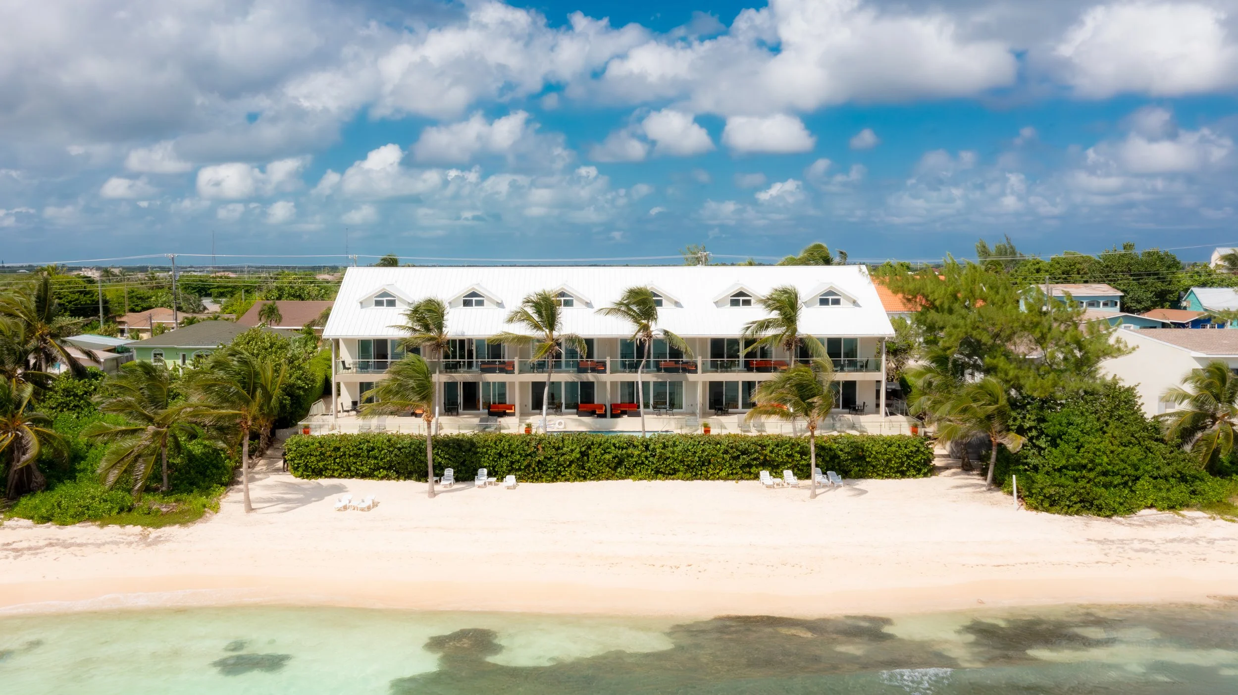 Oceanfront hotel with white exterior, balconies with orange accents, surrounded by palm trees, beach chairs, and sandy beach with clear water in the foreground, blue sky with scattered clouds.