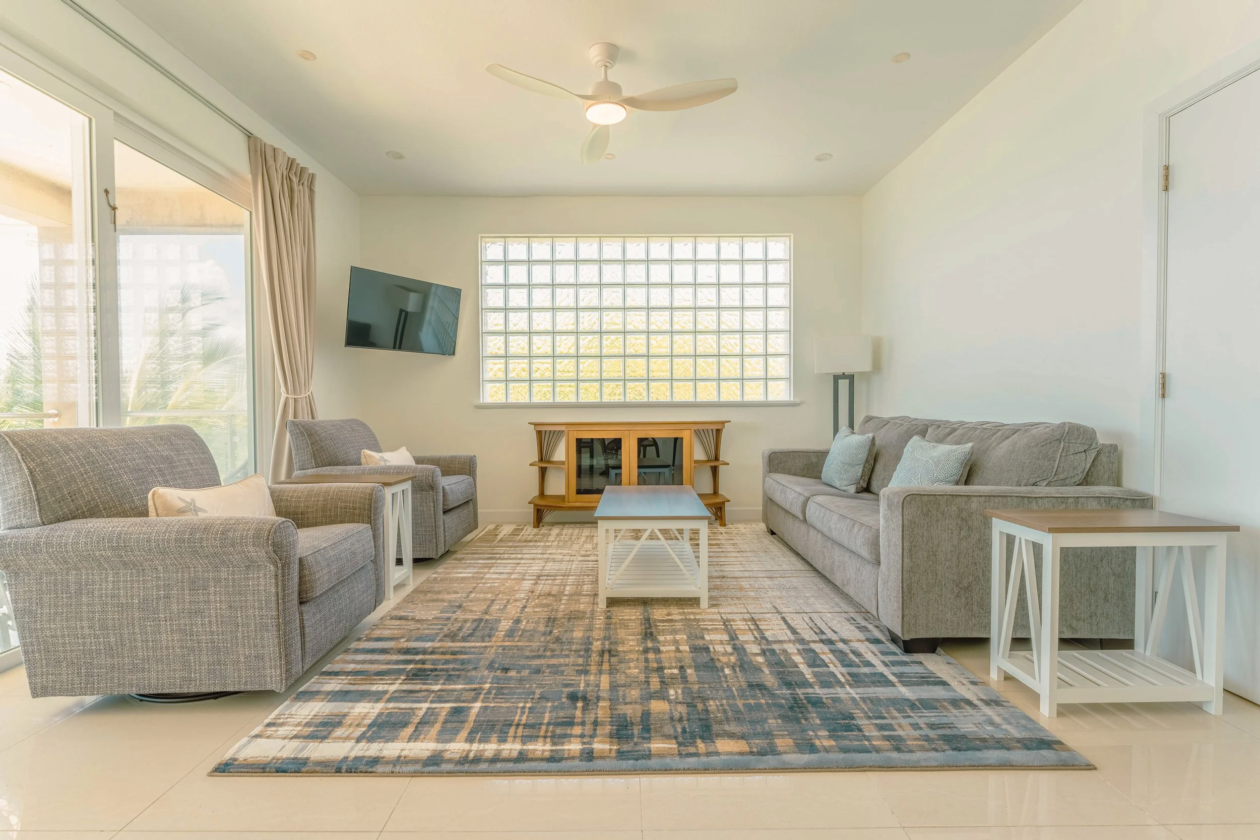 Living room with gray sofas, a wooden coffee table, a TV mounted on the wall, a floor lamp, a glass block window, beige curtains, and a ceiling fan.