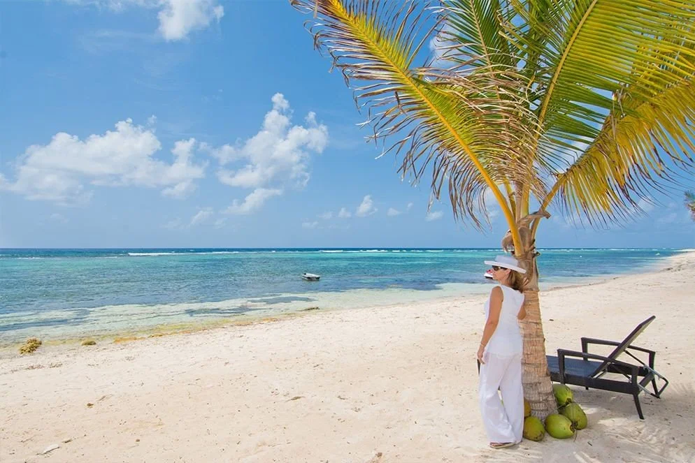 A woman standing next to a palm tree on a sandy beach, wearing white clothing, a sunhat, and sunglasses, with a few coconuts on the ground nearby, overlooking the ocean with a boat in the water under a partly cloudy sky.