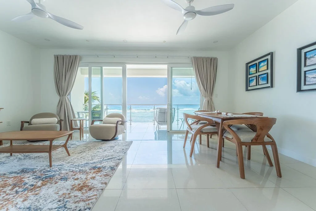 Living room with white walls and tiled floor, ocean view through sliding glass doors, sitting area with armchairs and a coffee table, dining area with a wooden table and chairs, framed seaside artwork on the walls, ceiling fans, and a balcony with outdoor furniture and palm trees.
