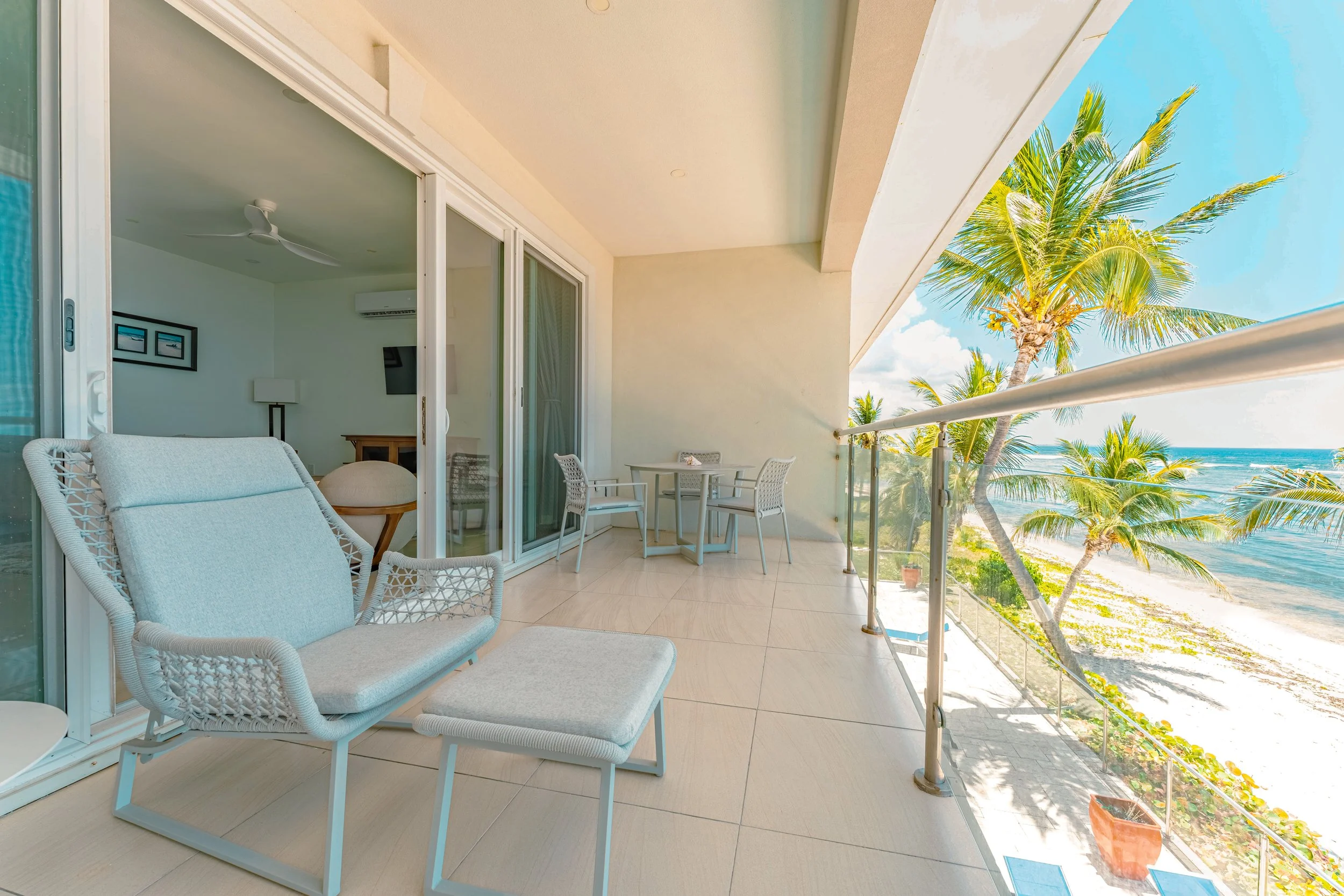 Balcony with white outdoor furniture, including a loveseat and a small table with two chairs, overlooking beach and palm trees.