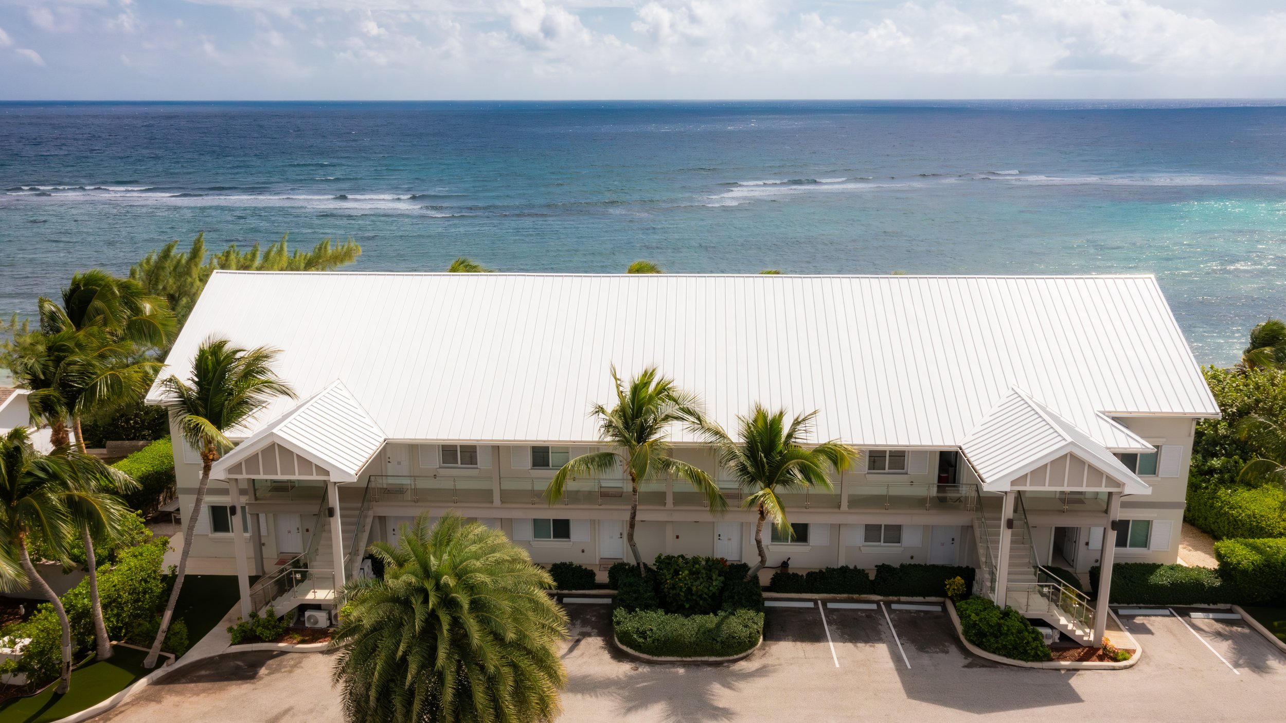 A white beachfront building with a metal roof, surrounded by palm trees, overlooking the ocean under partly cloudy skies.