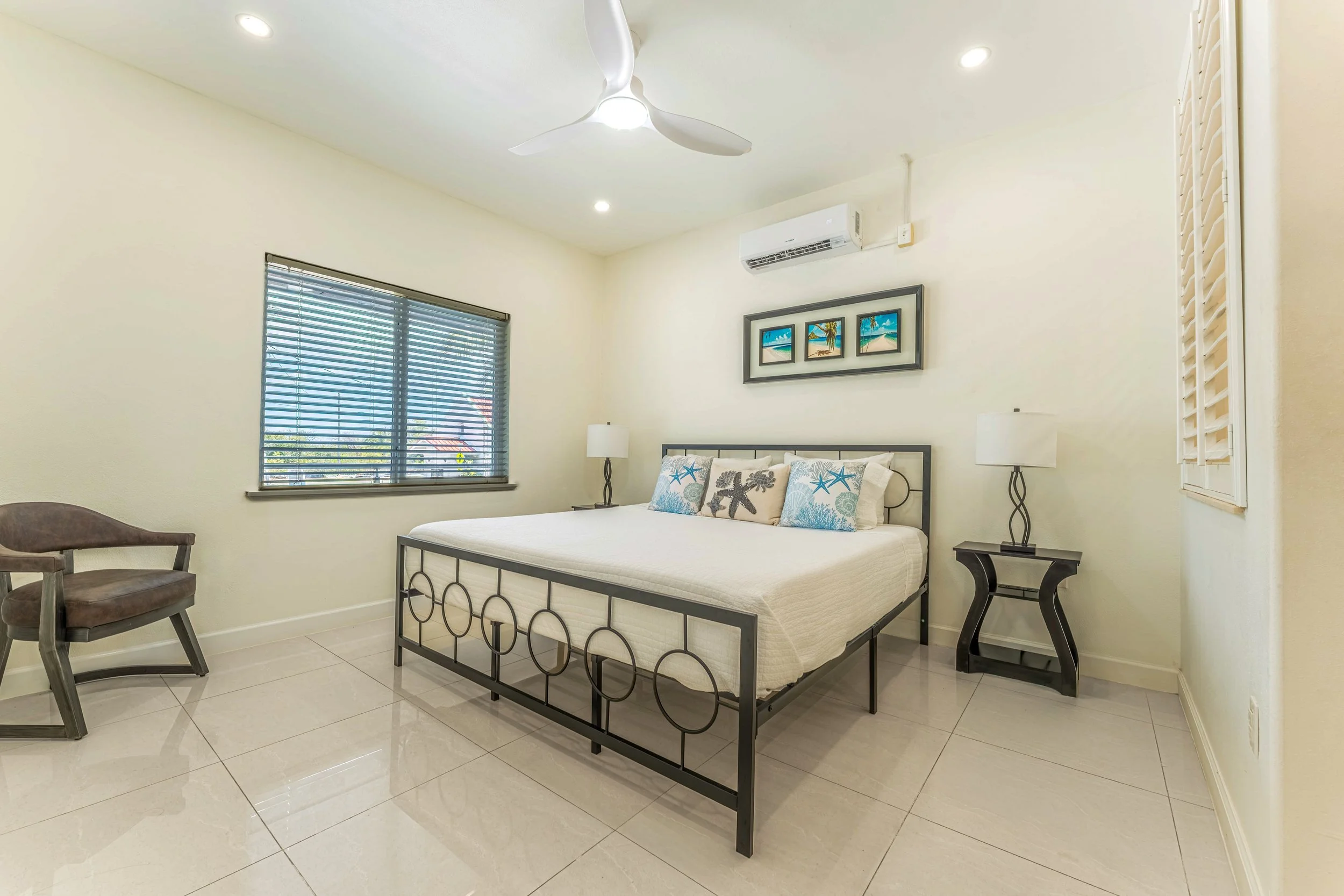 A bedroom with a white bed, decorative pillows with sea star and coral patterns, black metal bed frame, nightstands with white lamps, wall art depicting beach scenes, a window with blinds, and ceiling fan. The room has cream-colored walls and tiled f