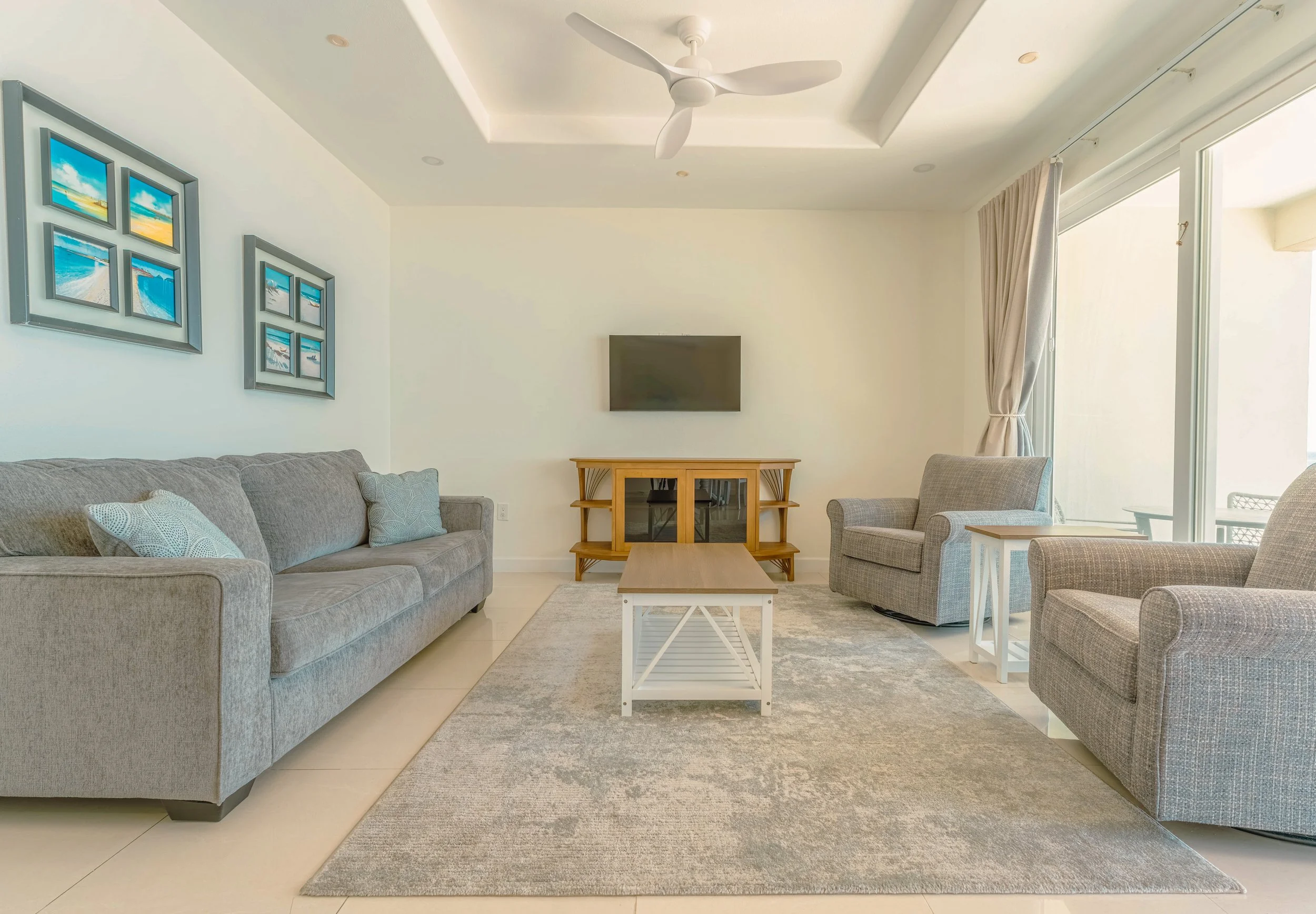 A modern living room with two gray sofas, a beige armchair, a wooden TV stand with a wall-mounted TV, a ceiling fan, framed beach-themed pictures on the wall, a large window with curtains, and a gray rug on a white tiled floor.