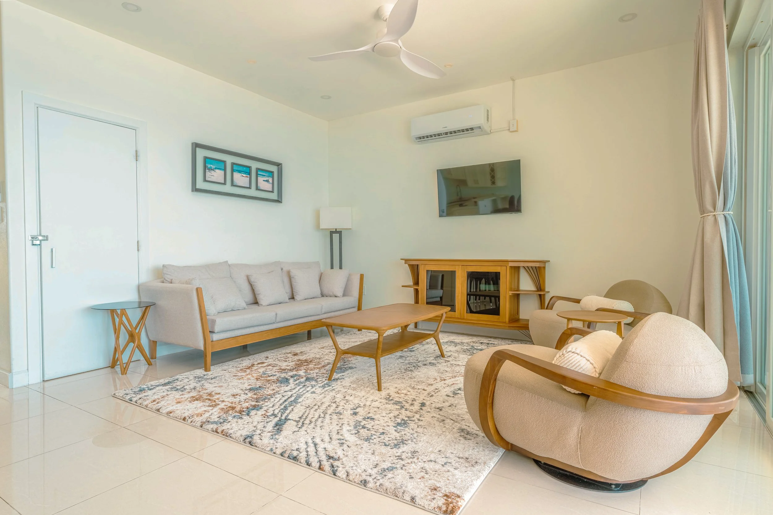 Living room with beige sofas, wooden furniture, a flat-screen TV, a ceiling fan, a display cabinet, a floor lamp, paintings on the wall, and large windows with beige curtains.