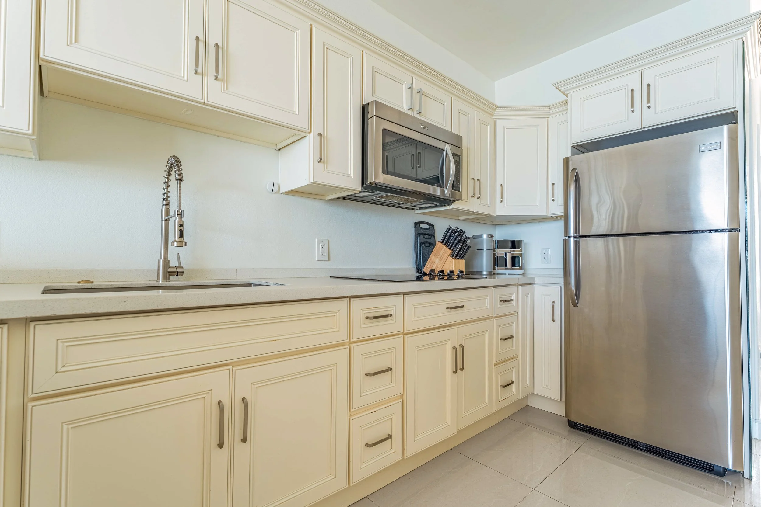 Kitchen with cream-colored cabinets, stainless steel microwave, and refrigerator, with a sink and faucet, black knife set, and small kitchen appliances on the counter.