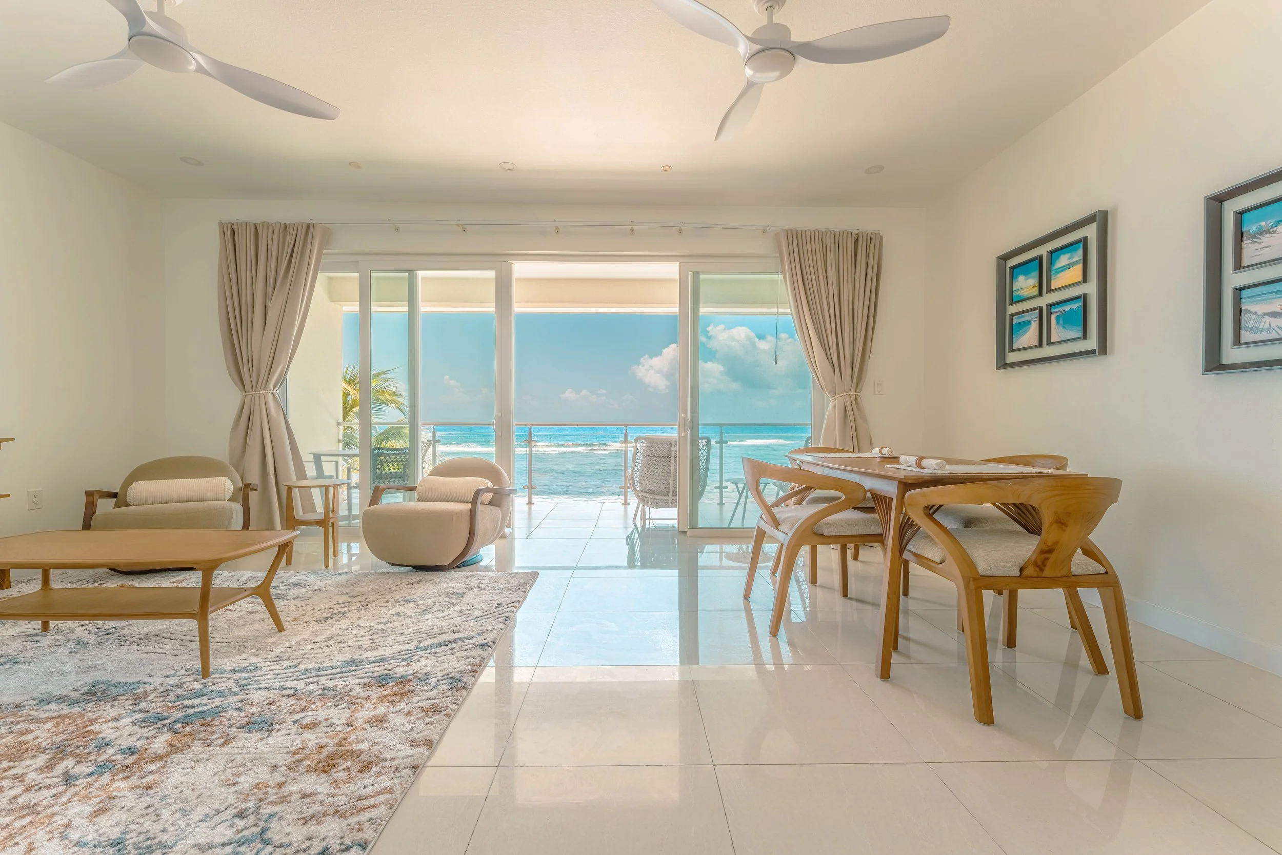 Living room with large glass doors leading to a beach view, beige furniture, a patterned area rug, framed beach artwork, and ceiling fans.