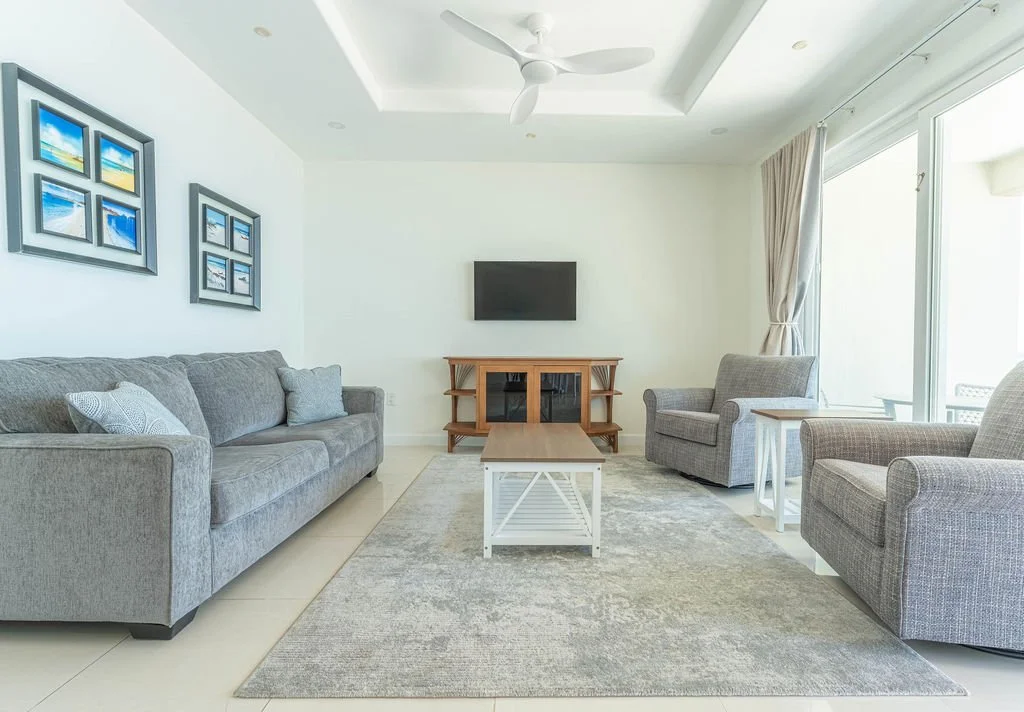 Modern living room with light gray sofa, two matching armchairs, wall-mounted flat-screen TV, small wooden TV stand, white coffee table, side table, artwork on wall, large sliding glass door with curtains, ceiling fan, neutral-toned rug.