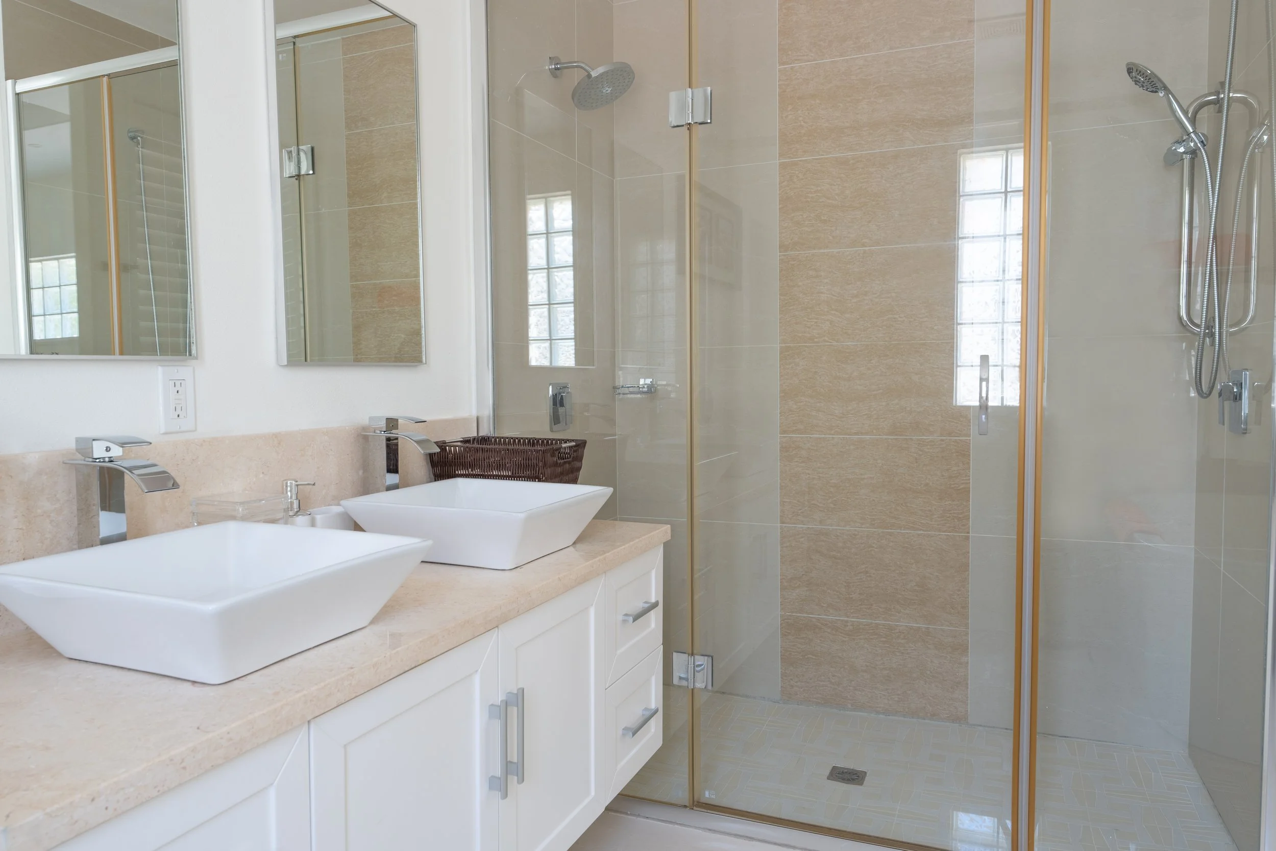 A modern bathroom with a double sink vanity, large mirrors, a glass-enclosed shower with rainfall and handheld showerheads, beige tiled walls, and a window providing natural light.