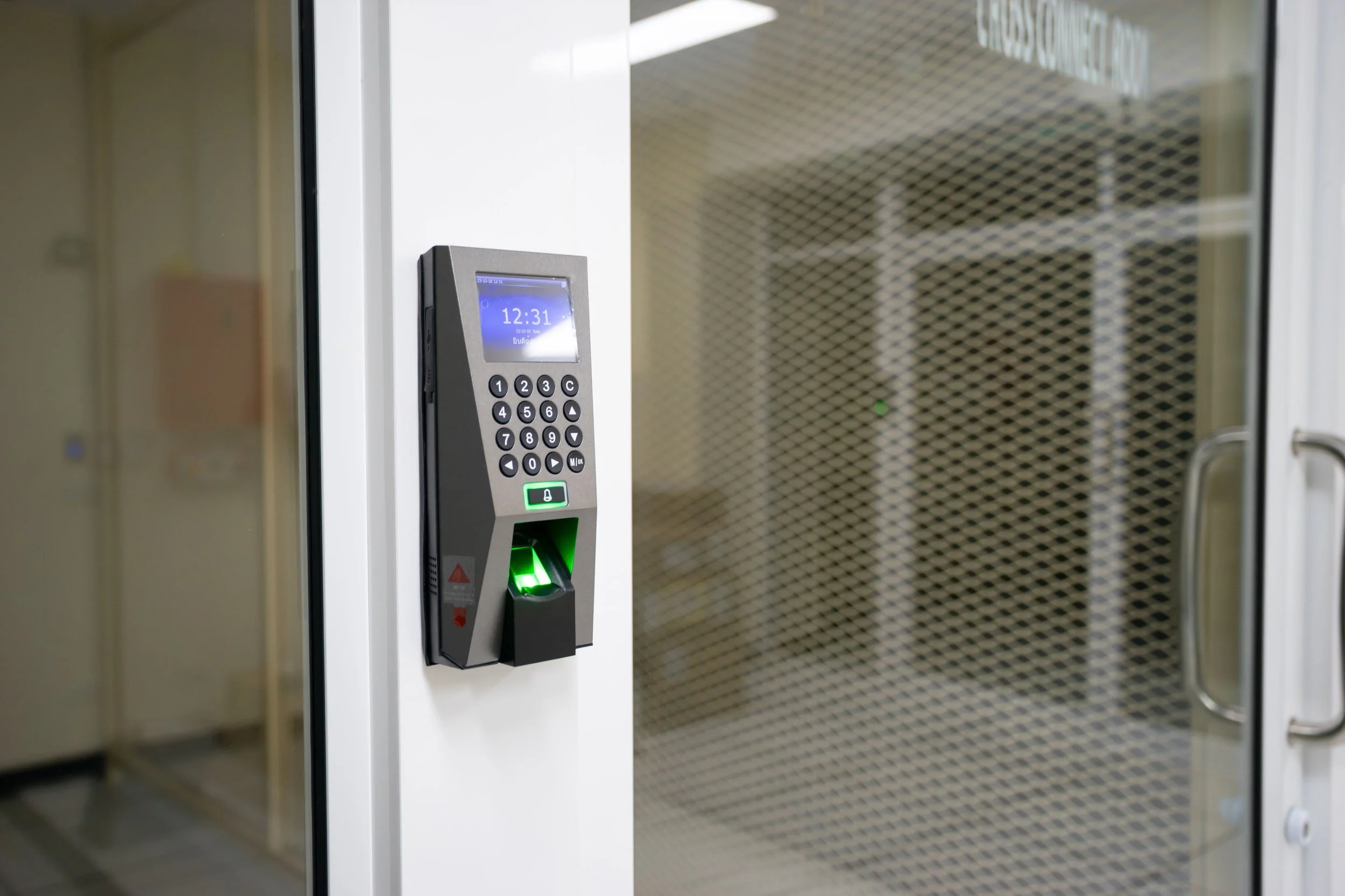 Foto di un sistema di accesso con riconoscimento biometrico a vuoto, con un display digitale che mostra l'ora 12:31 e un codice. Il sistema è installato sulla porta di vetro di un edificio, con una maniglia sulla destra.