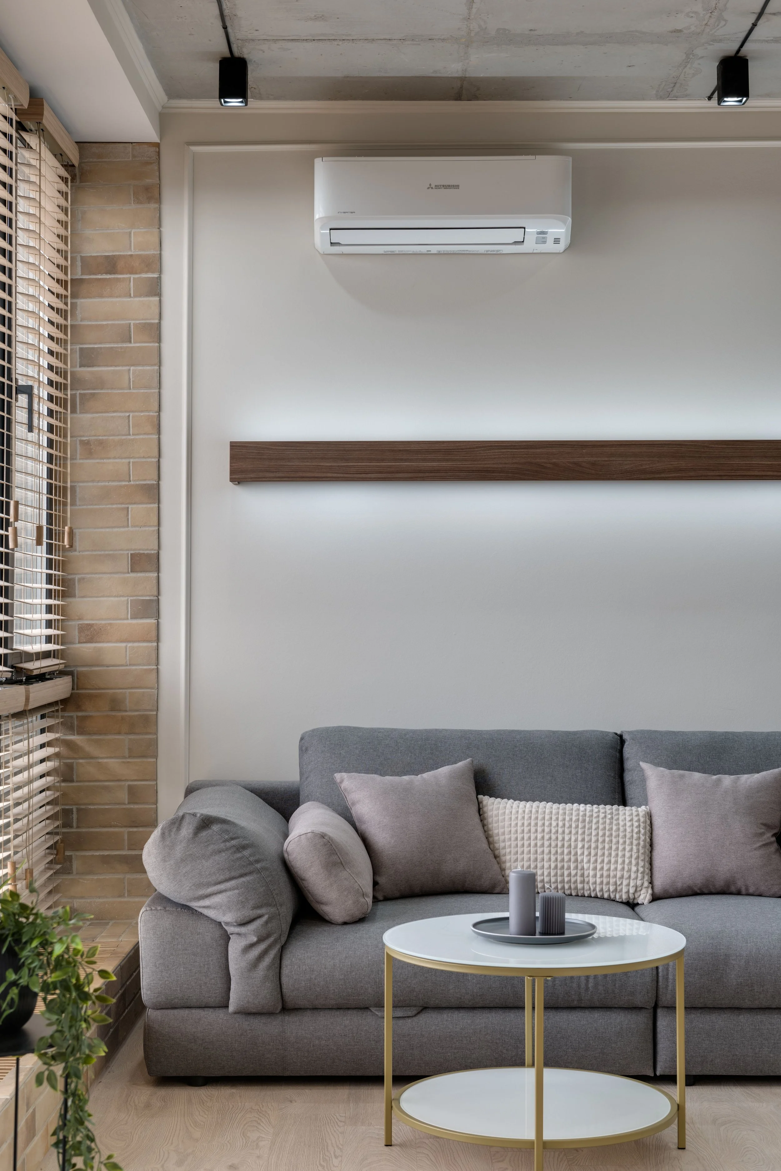 Soggiorno moderno con divano grigio e cuscini, tavolino circolare bianco con struttura dorata, aria condizionata bianca, pareti chiare, porta di vetro con persiane di legno, elementi decorativi minimalisti.