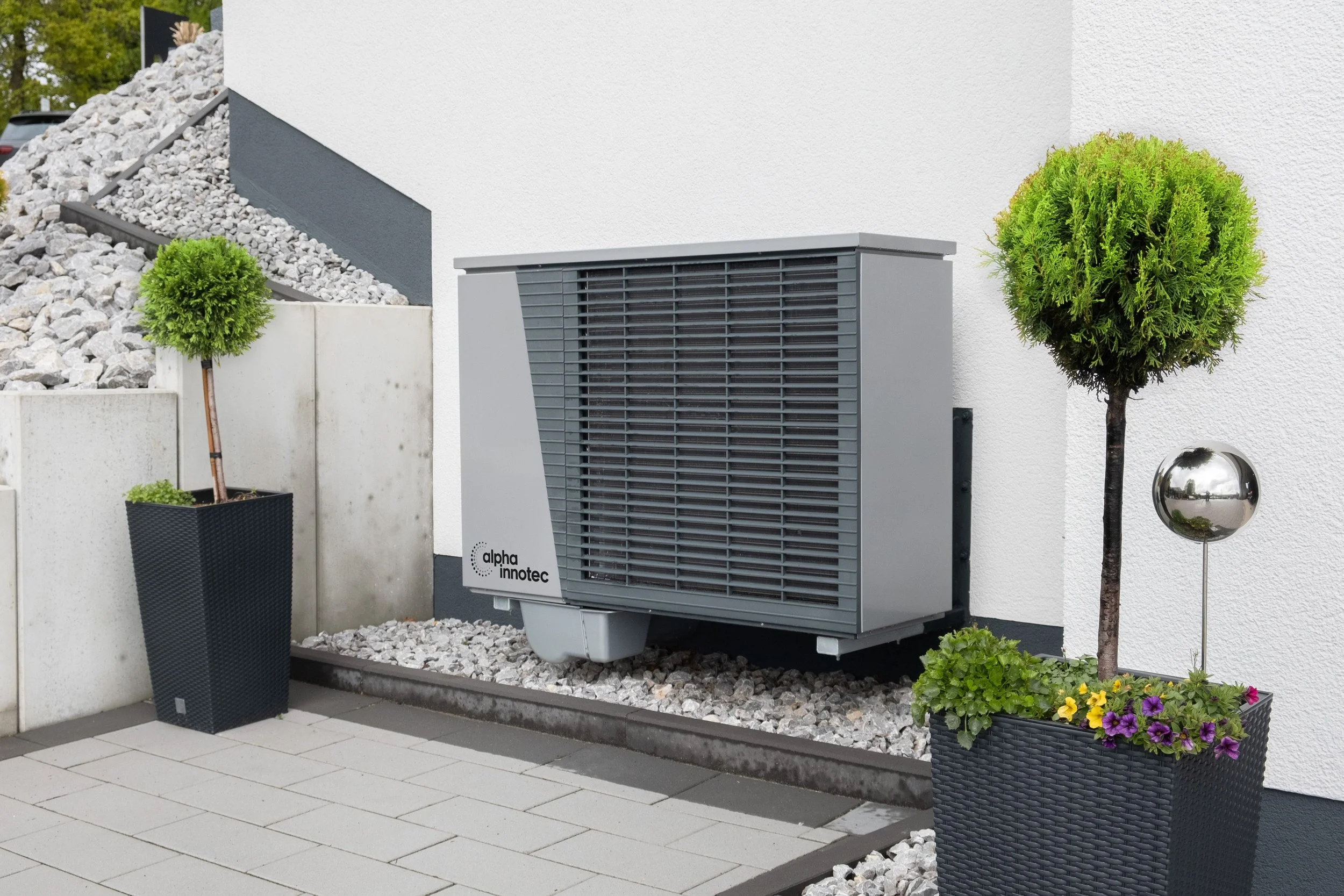 Esterno di un edificio moderno con un'unità di condizionamento Alpha Innotec, piante ornamentali e piante in ciotole nere.