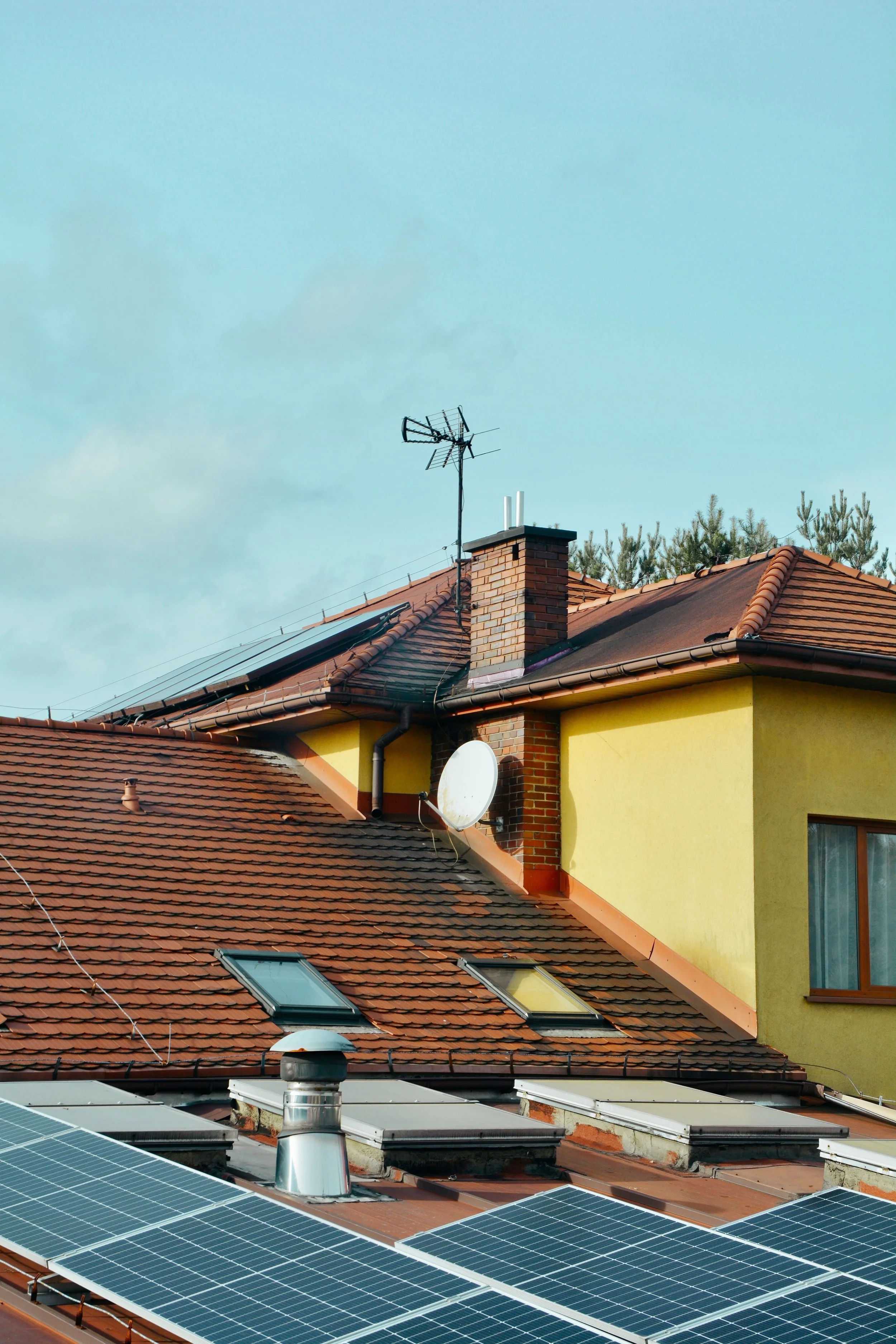 Tetto di una casa con pannelli solari e finestre panoramiche, camino e antenna TV, con tetto in tegole rosse e pareti gialle, con alberi sullo sfondo e un cielo azzurro