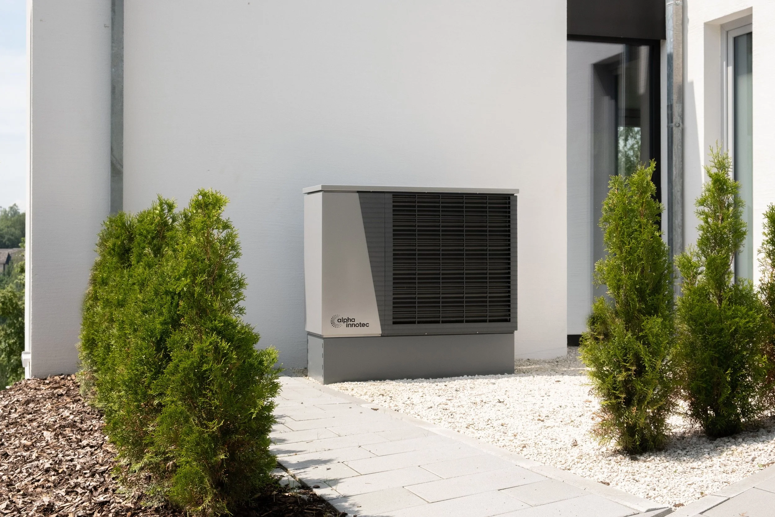 Climatizzatore esterno Alfa Innotec installato su terrazza di una moderna abitazione con parete bianca e vegetazione verde.