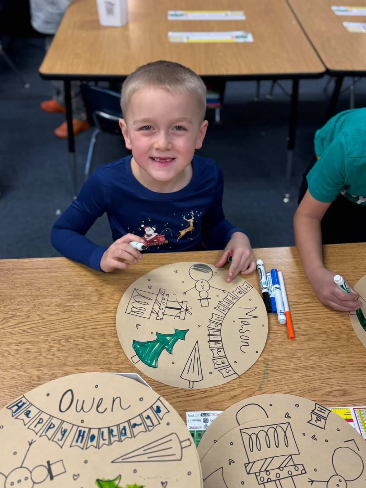 A young boy with short blonde hair smiling at the camera while sitting at a classroom table. In front of him is a round piece of paper with Christmas-themed drawings and the name "Mason" written on it. The paper includes a Christmas tree, gifts, a snowman, and the words "Merry Christmas." There are markers on the table, and other children's papers are partially visible.