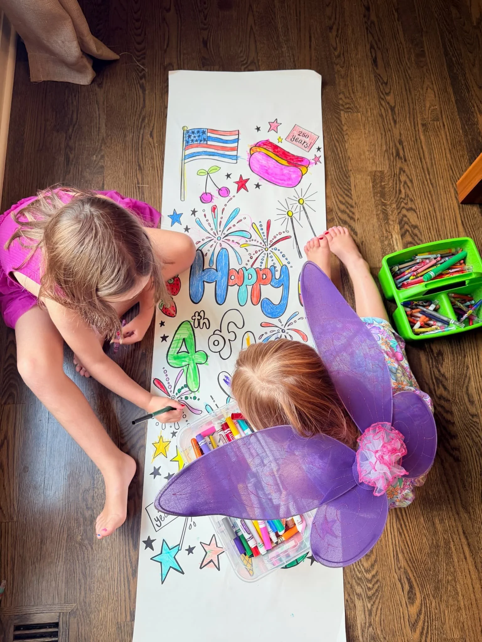 Children sitting on a wooden floor coloring a large poster with patriotic and celebratory drawings, including an American flag, fireworks, and a hot dog. There is a green container filled with colorful markers nearby.